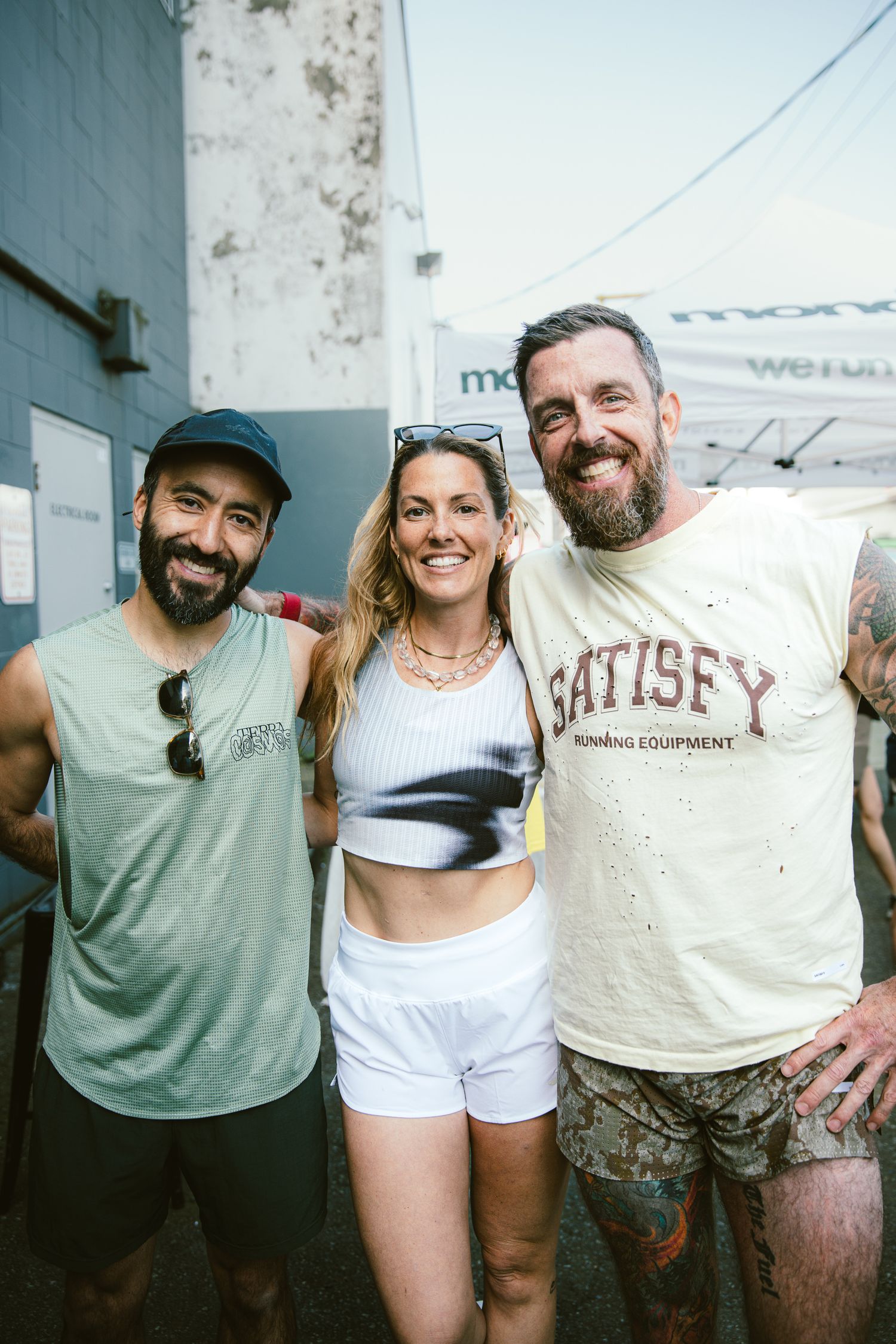 Three smiling friends pose together outdoors wearing Satisfy Running Equipment apparel after a race.
