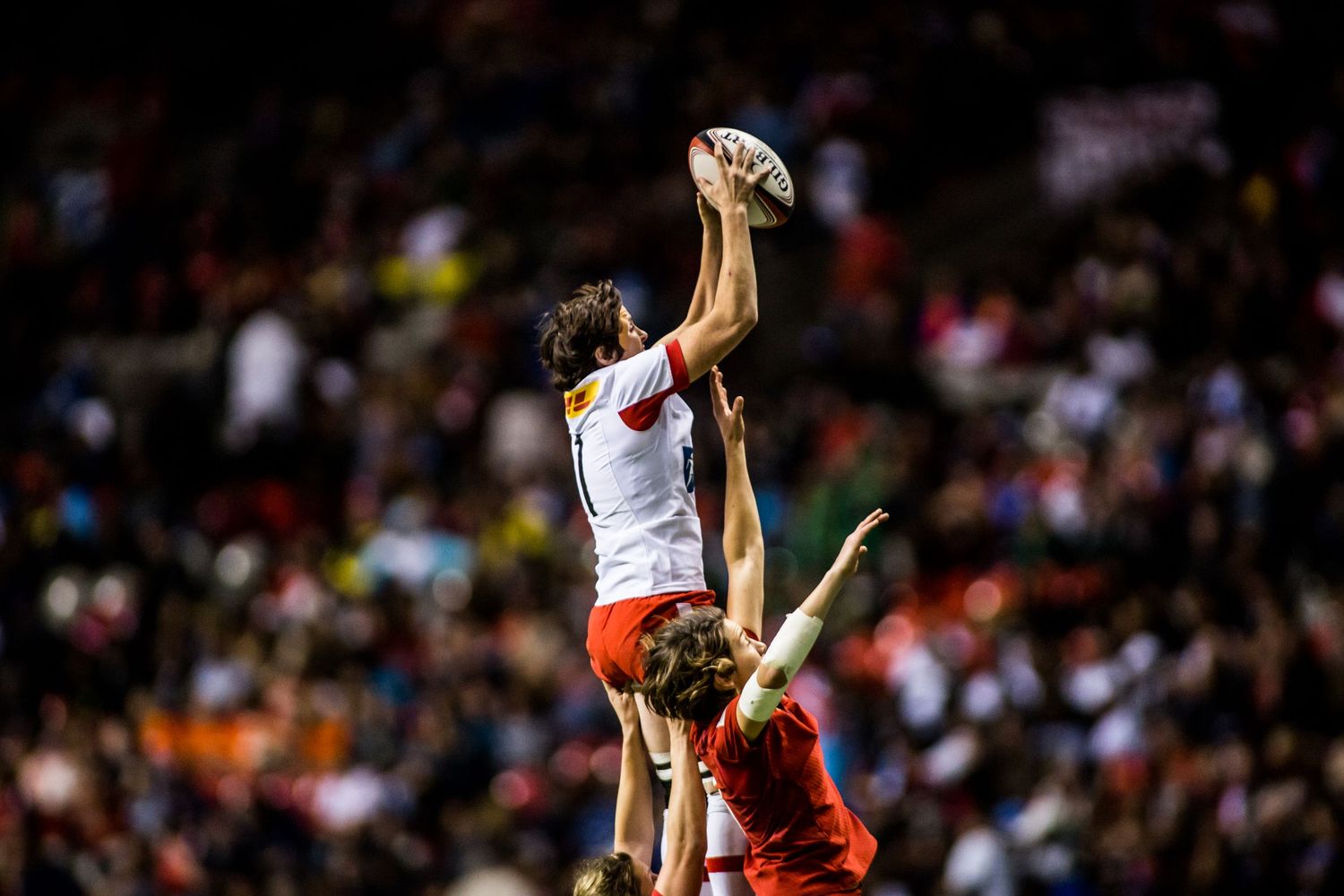 Rugby player leaps high to catch ball in lineout during a competitive night match with crowd in background.