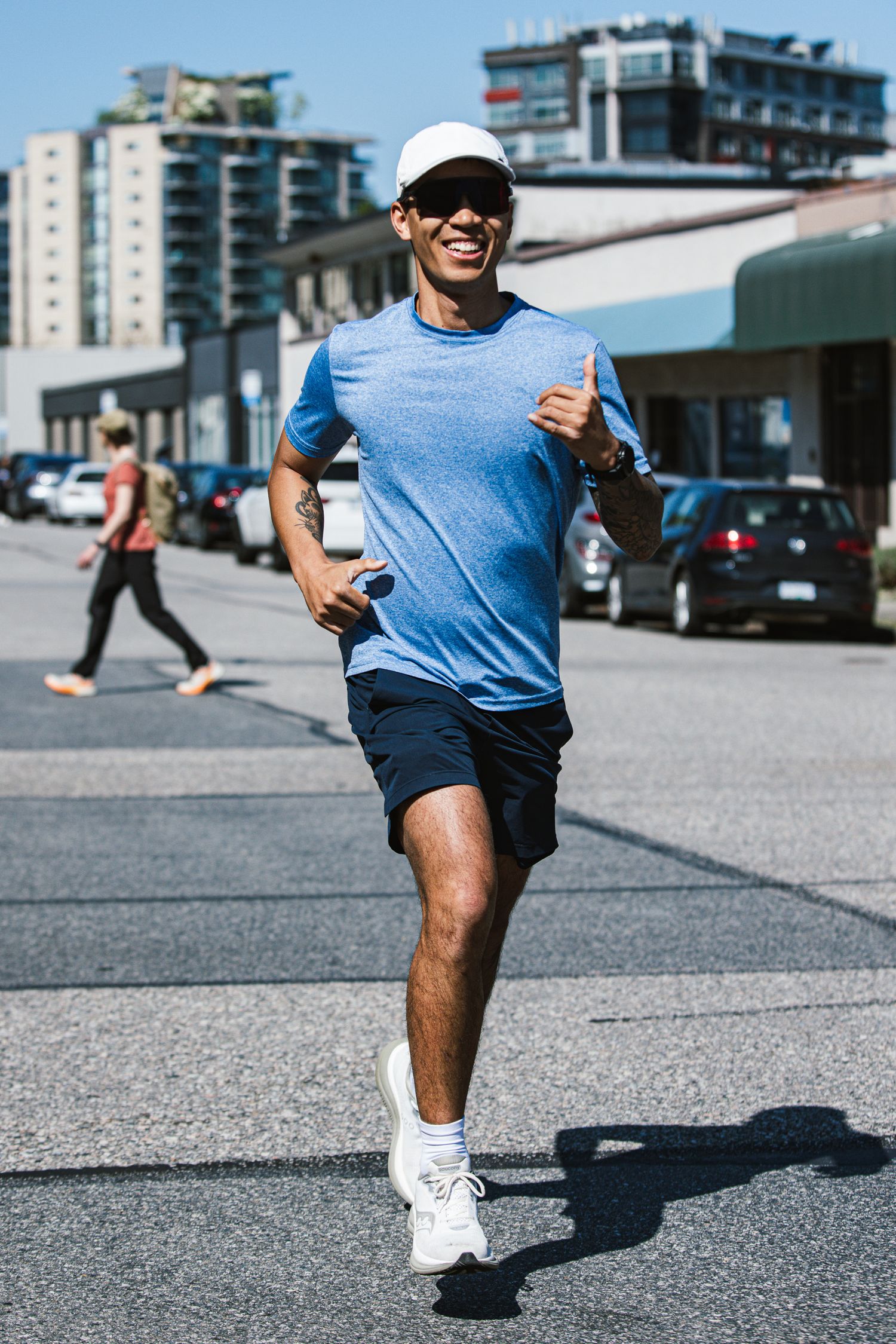 Smiling man in blue t-shirt, white cap, and navy shorts jogging on a sunny urban street.