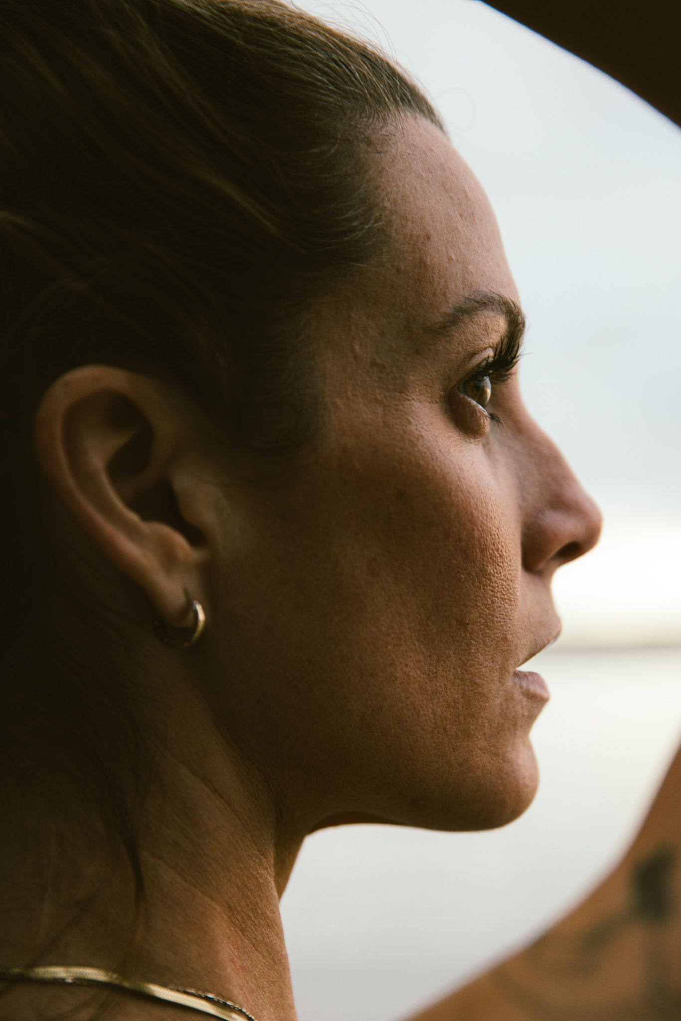 Side profile of athletic woman with gold hoop earring and necklace in natural light.