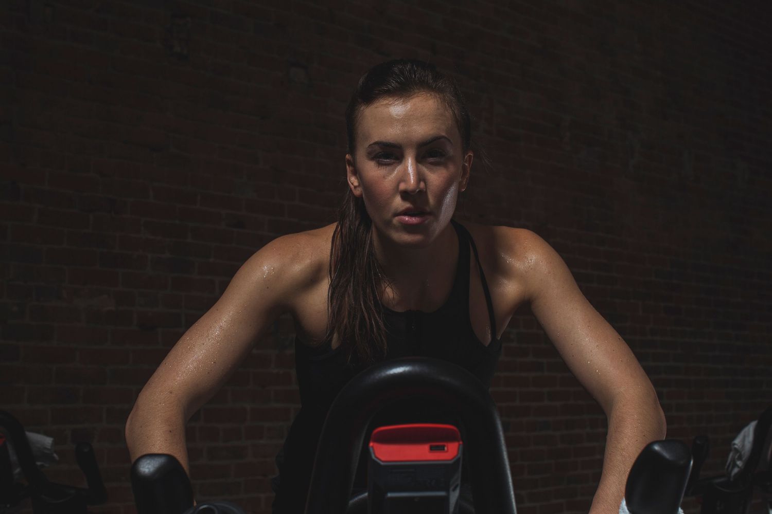 Sweaty woman riding stationary bike during intense indoor cycling workout in dark gym.