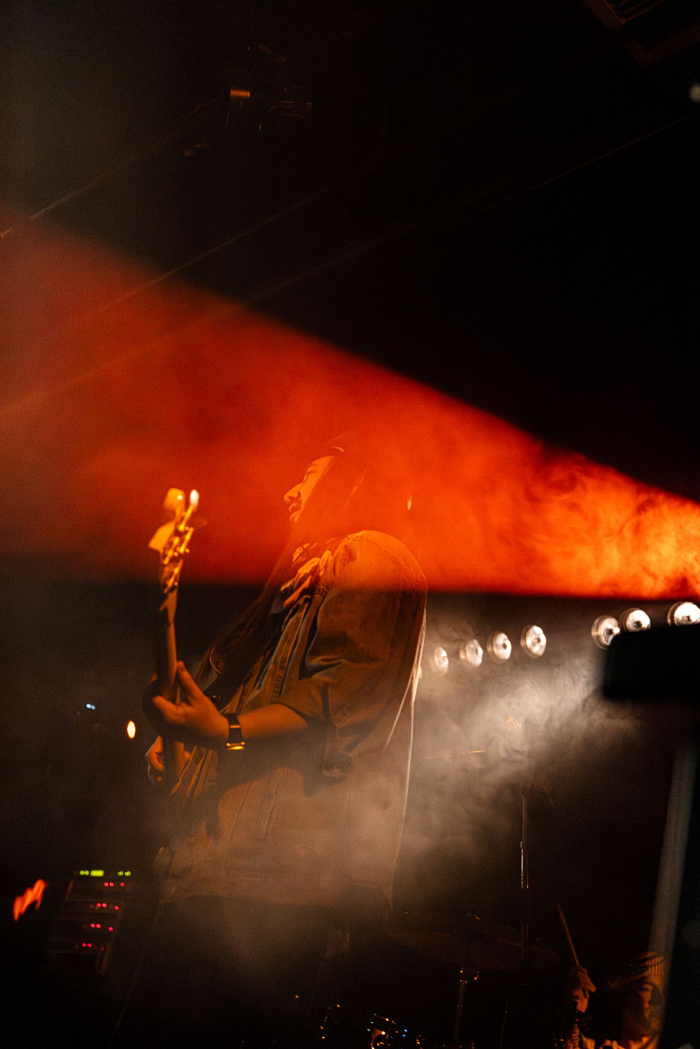 Guitarist performs on a dramatic smoke-filled stage illuminated by an intense red and orange spotlight beam.