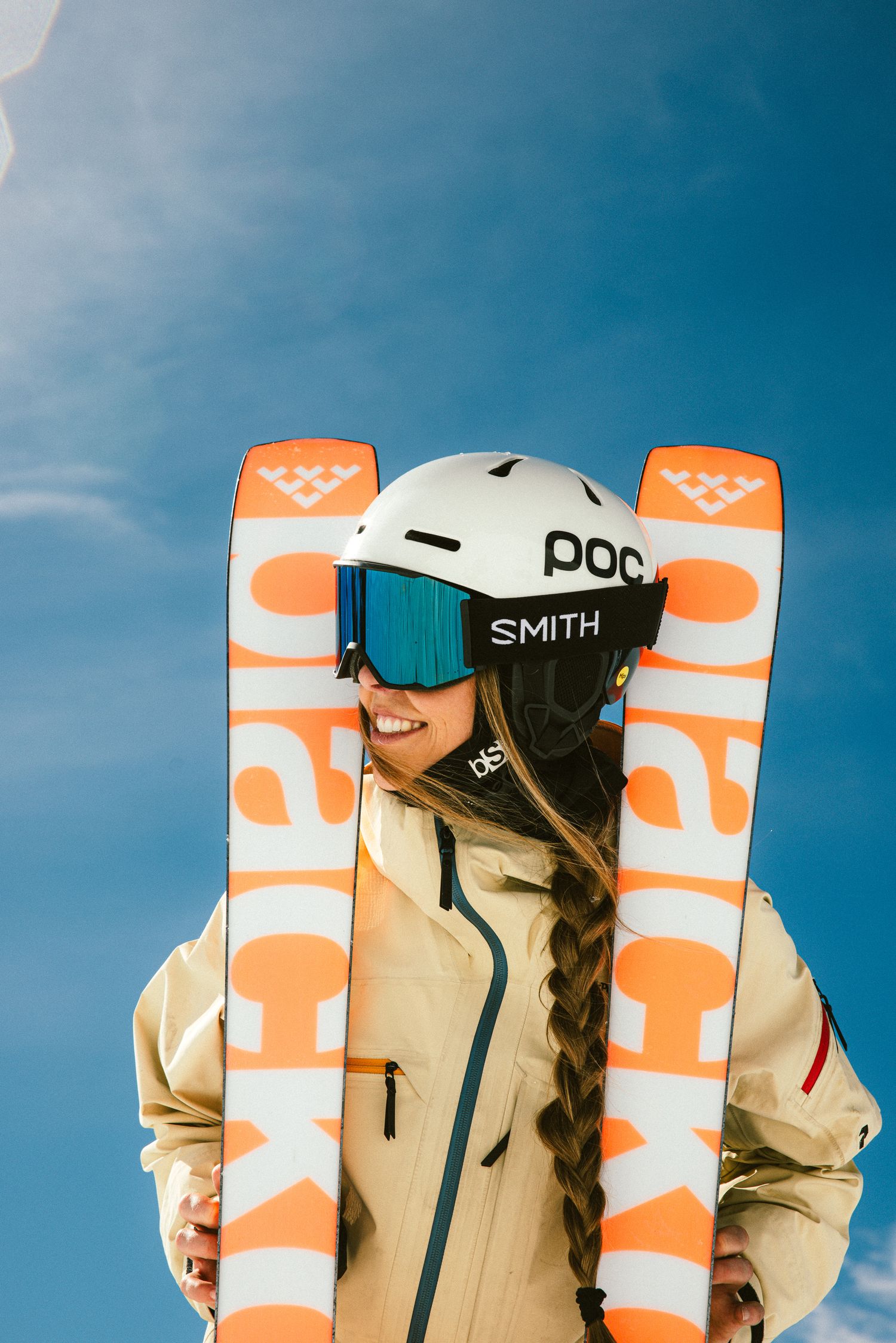 Female skier smiling with orange Black Crows skis framing her face wearing POC helmet and Smith goggles under blue sky.