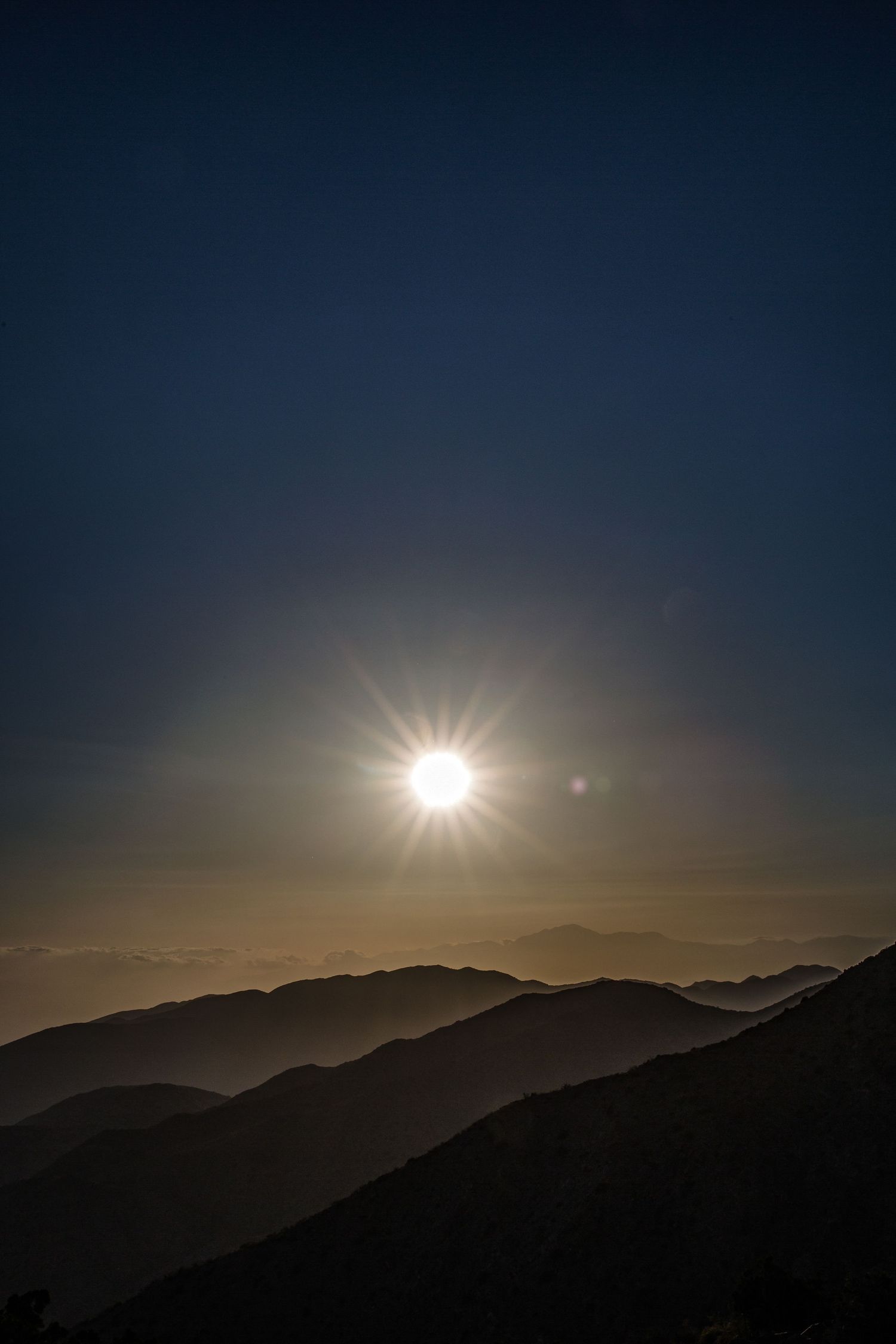 Bright sun star setting over misty layered mountain ridges under a deep blue sky at dusk.