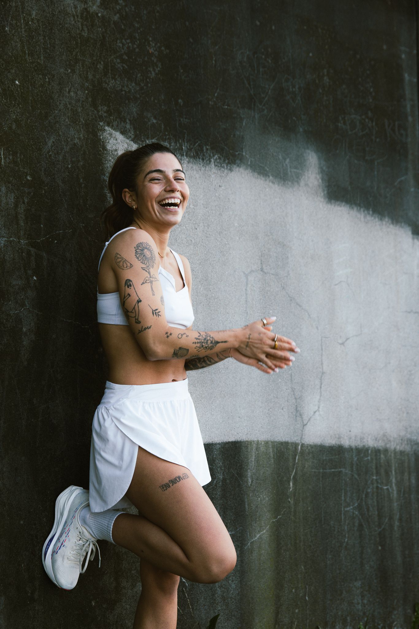 Laughing tattooed woman in white athletic outfit leaning against a dark concrete wall outdoors.