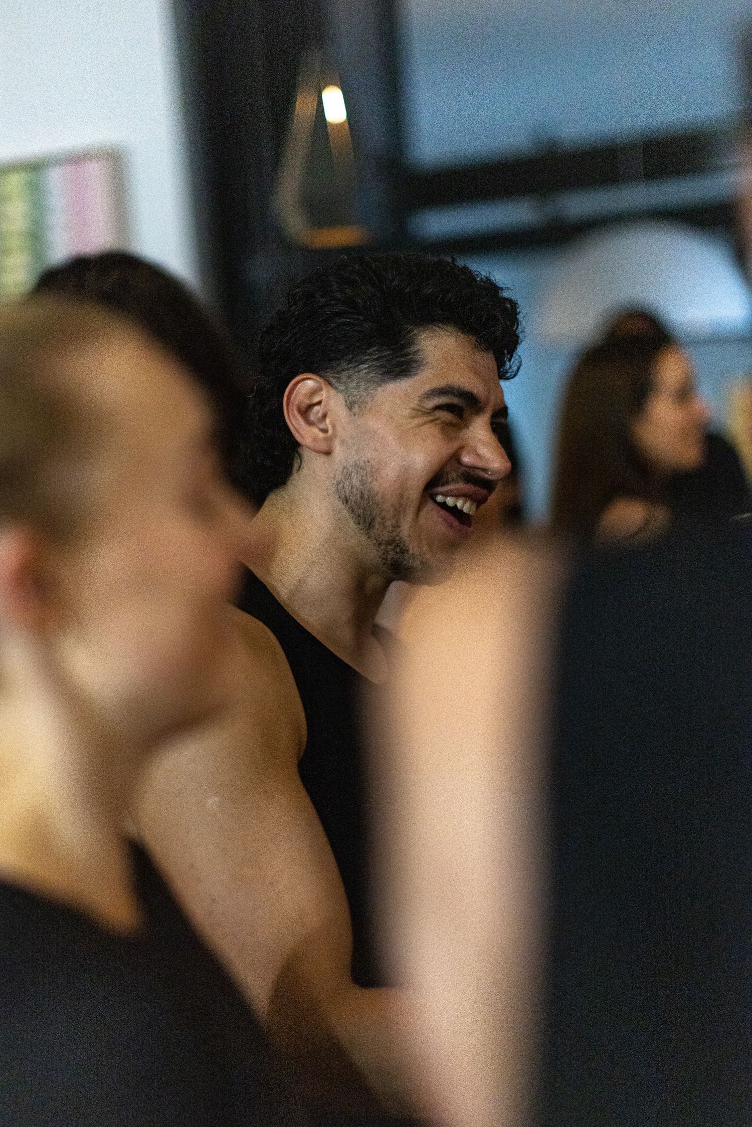 A muscular man with curly hair laughs joyfully in a gym setting, wearing a black tank top.