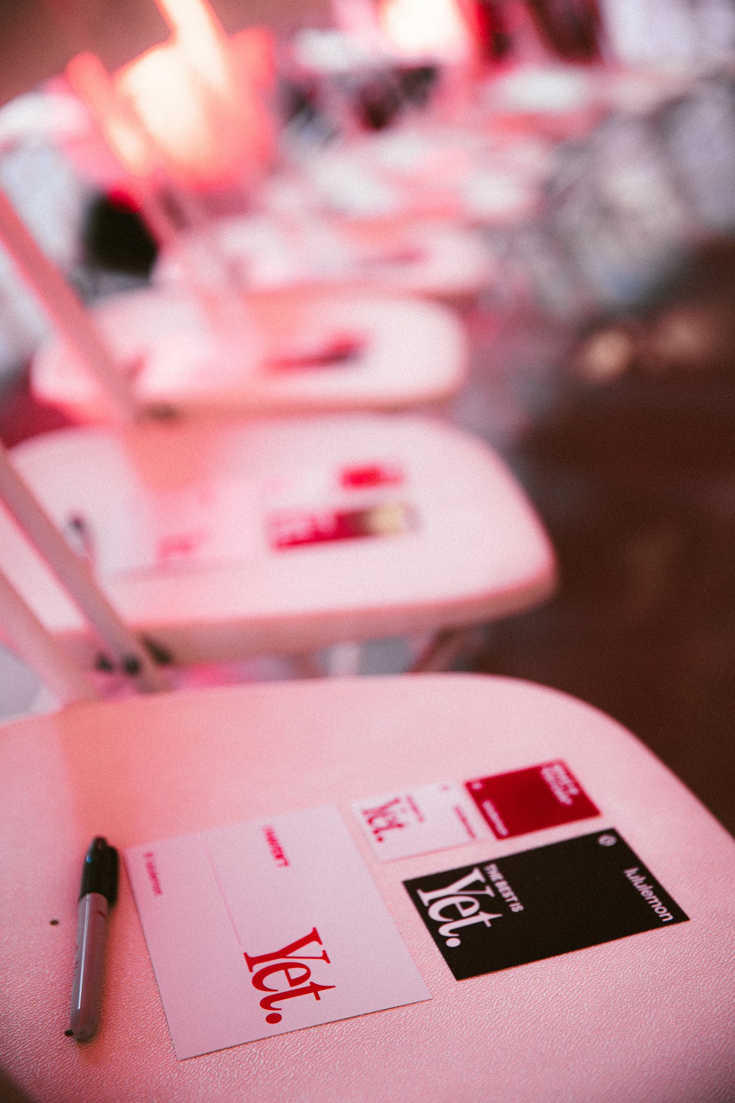 White folding chairs with red and black event cards and a pen under pink ambient lighting at a formal gathering.