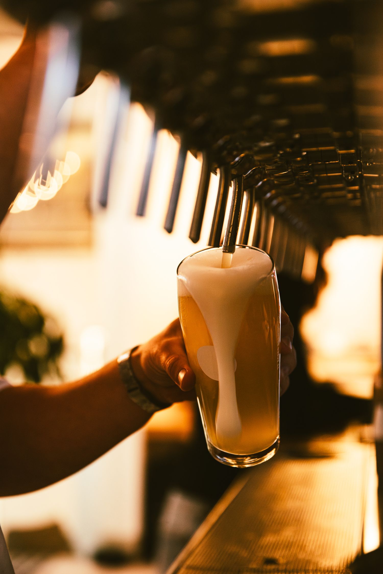 Bartender pouring a frothy draft beer from tap handles at a warmly lit bar.