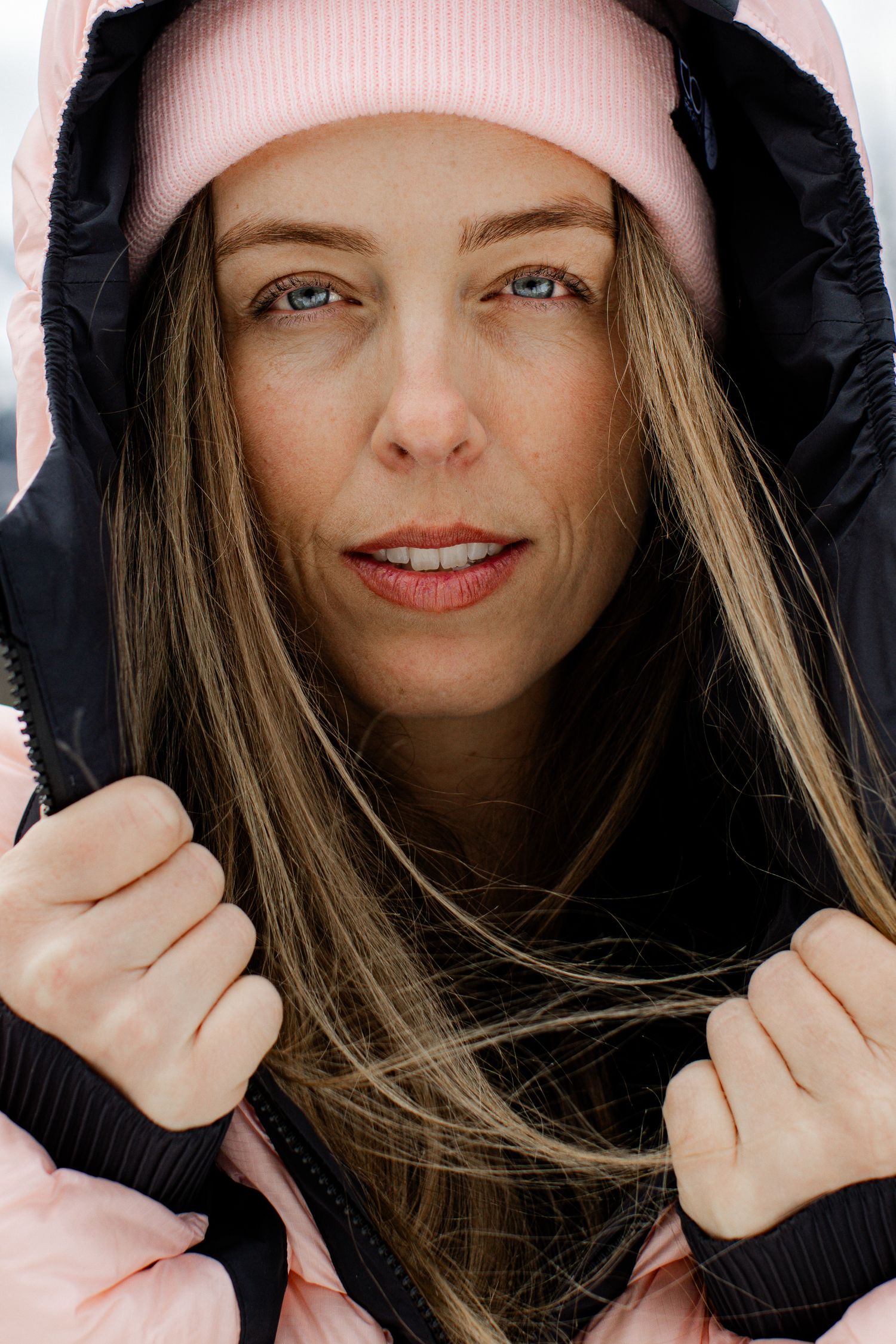Close-up portrait of blue-eyed woman wearing pink beanie and black hooded pink puffer jacket in winter setting.