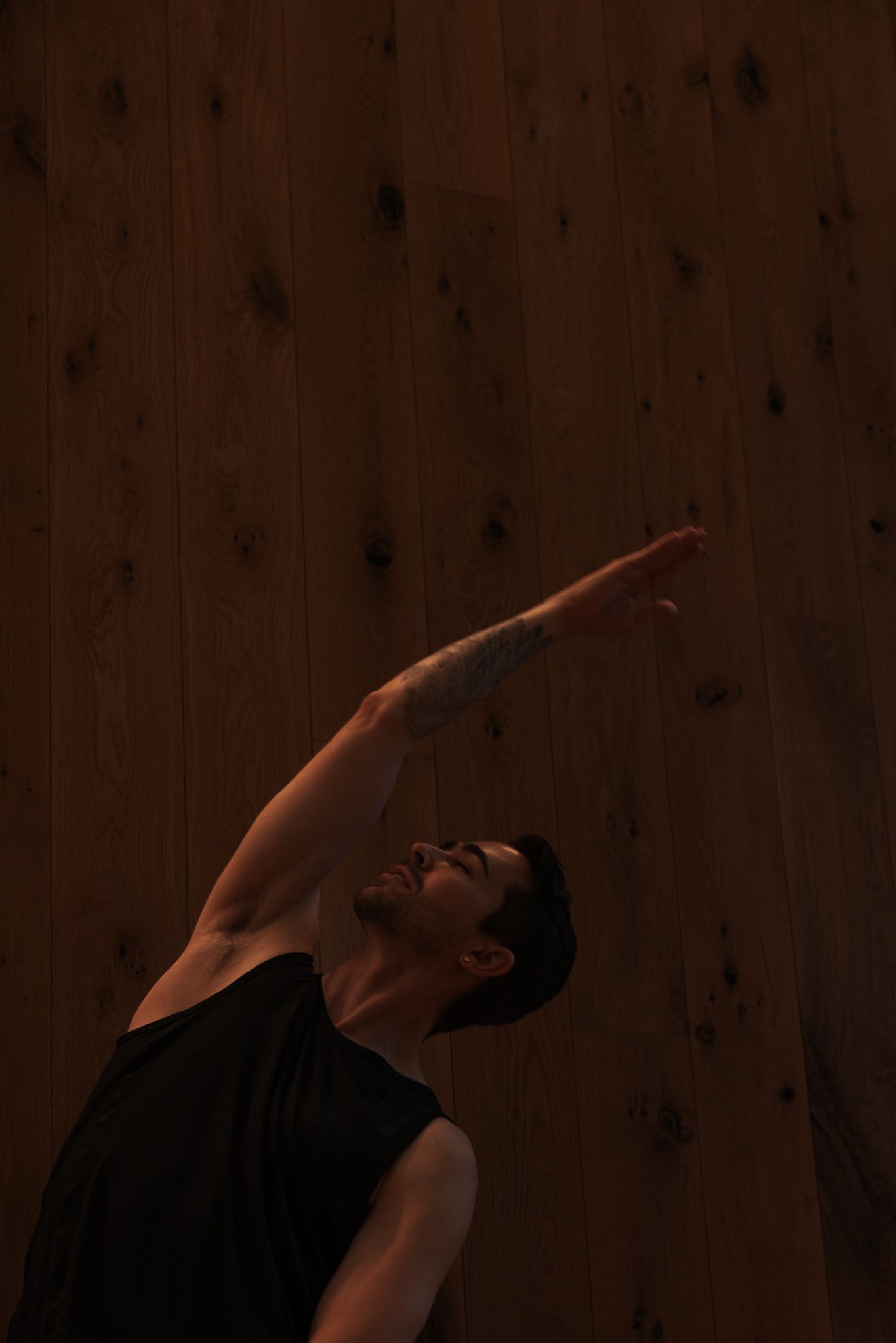 Person in black tank top performing yoga side stretch pose against dark wooden wall background.