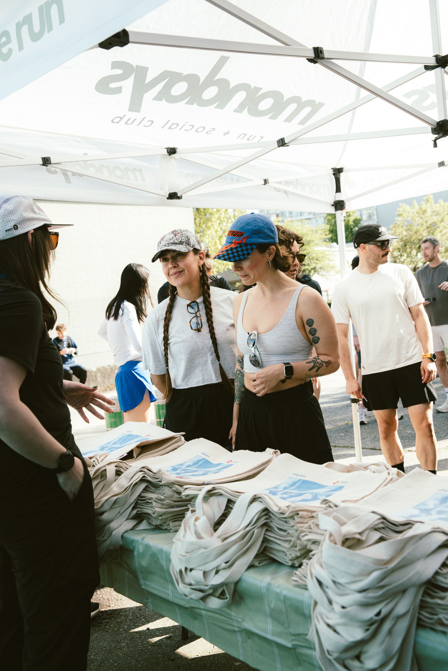 Two women browse merchandise at an outdoor event booth under a Mondays Run Club tent on a sunny day.
