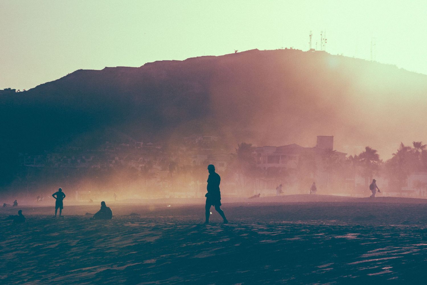 Silhouettes of people playing on a misty beach at sunset with a hill and city skyline in the background.