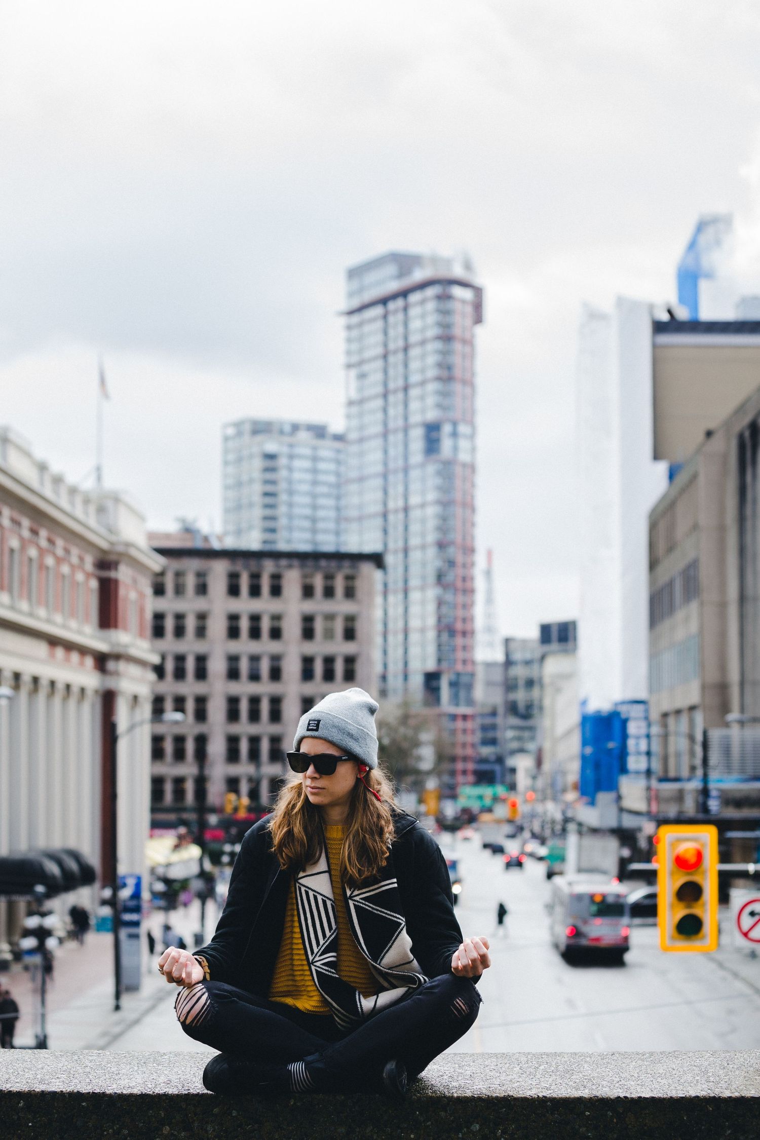 Woman meditating in lotus pose on urban ledge, wearing beanie and sunglasses, with city skyscrapers in background.