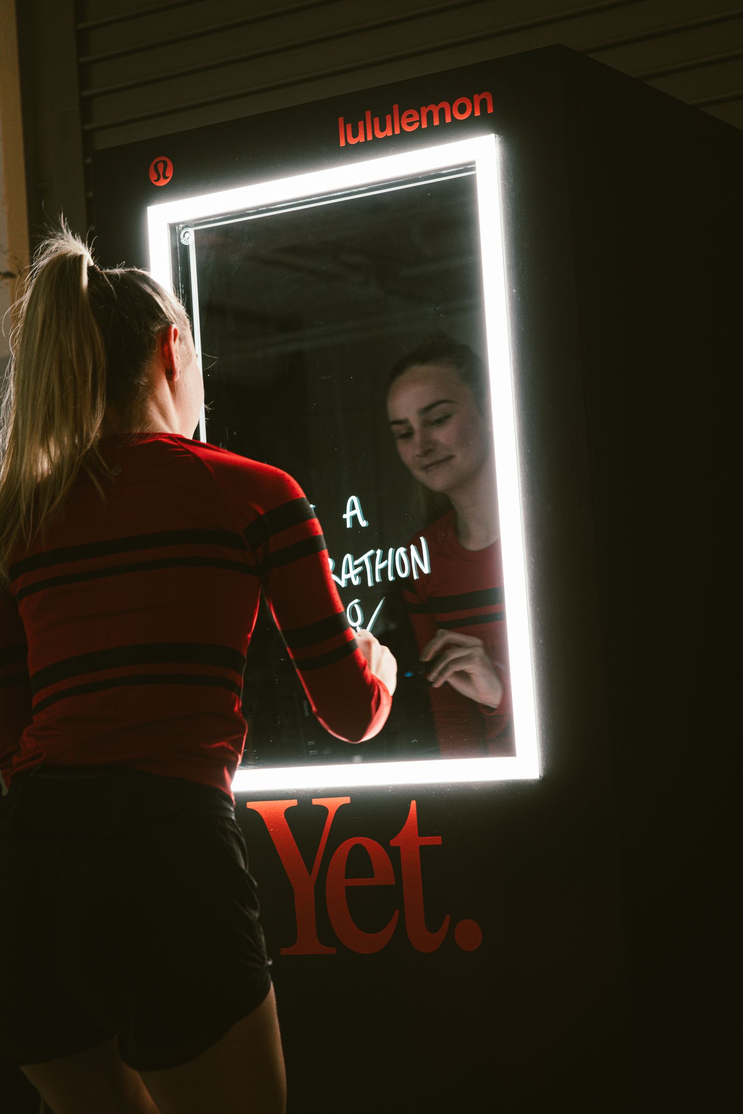 Woman writing on an illuminated lululemon Yet. interactive mirror display at a fitness event.