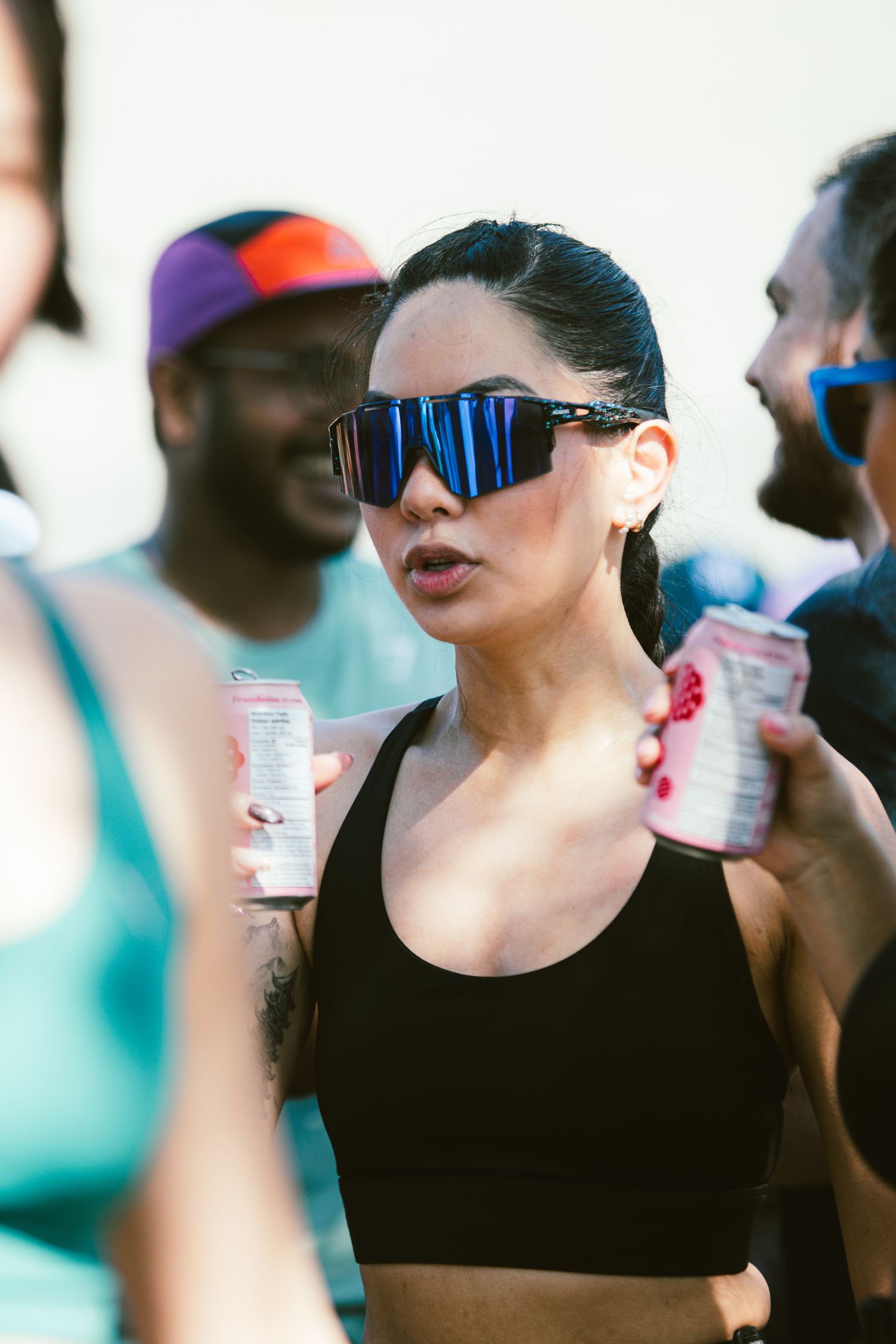 Woman in black sports bra and blue mirrored sunglasses holding cans at an outdoor fitness event.