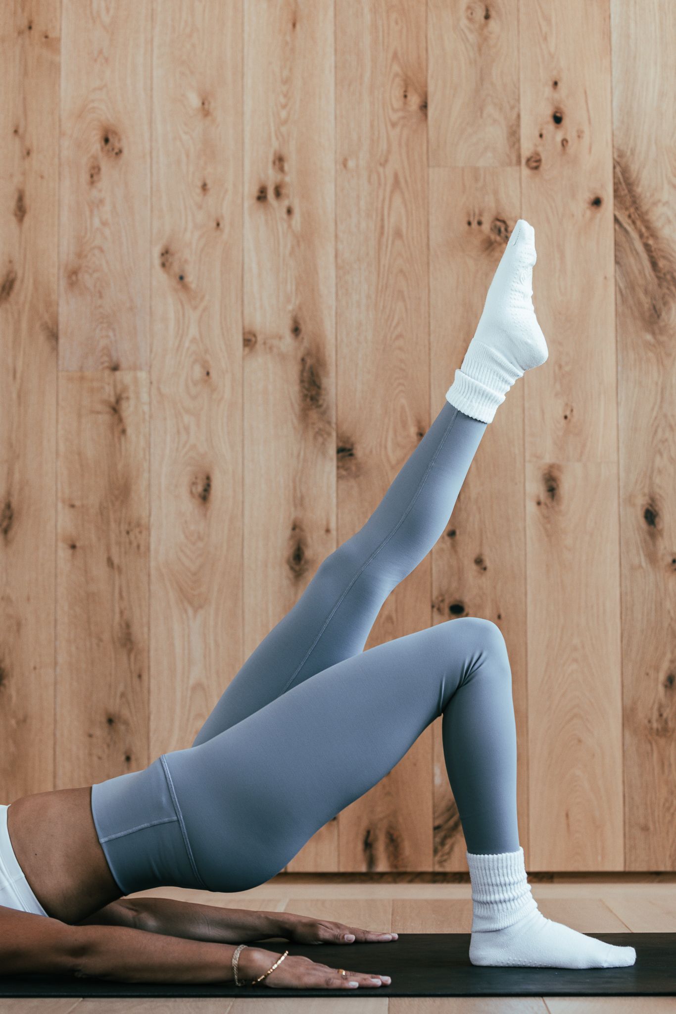 Woman in grey leggings and white socks performs a single-leg glute bridge on a black yoga mat.