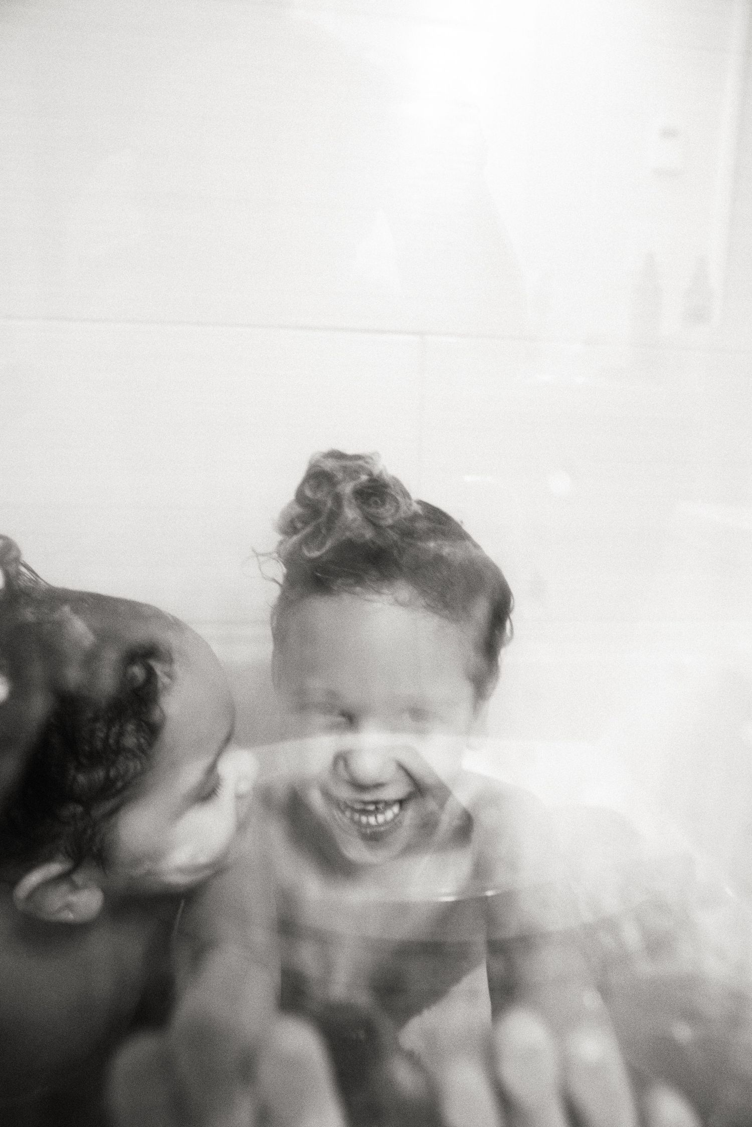 Black and white photo of two children laughing and playing in a bathtub viewed through wet glass.