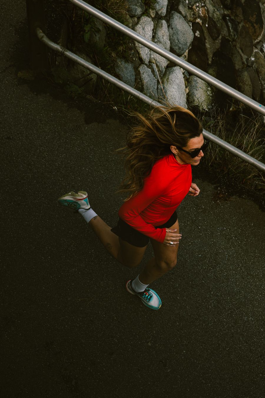Aerial view of a woman running in a red long-sleeve top, black shorts, and teal sneakers on a path.