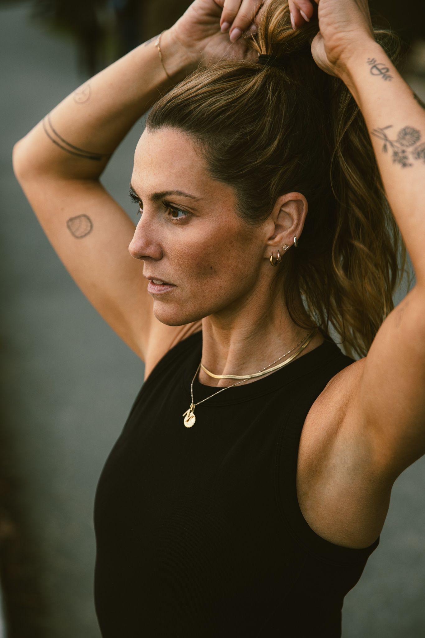 Fit woman with tattoos adjusting ponytail wearing black tank top and gold necklace.
