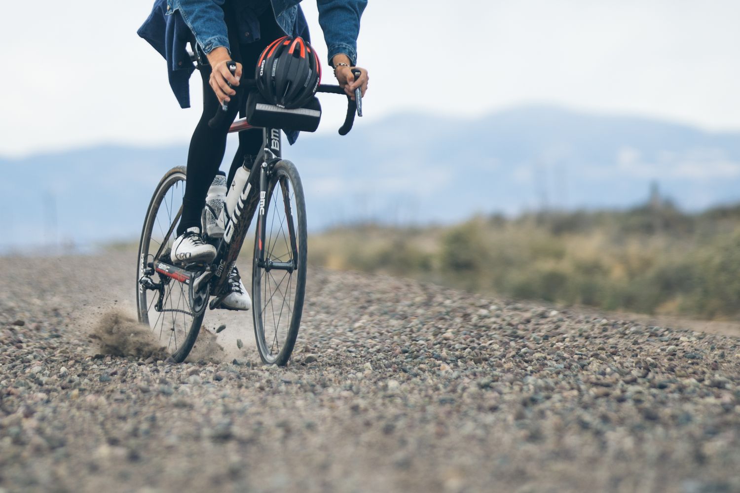 A cyclist rides a road bike on a gravel path with mountains and desert landscape in the background.