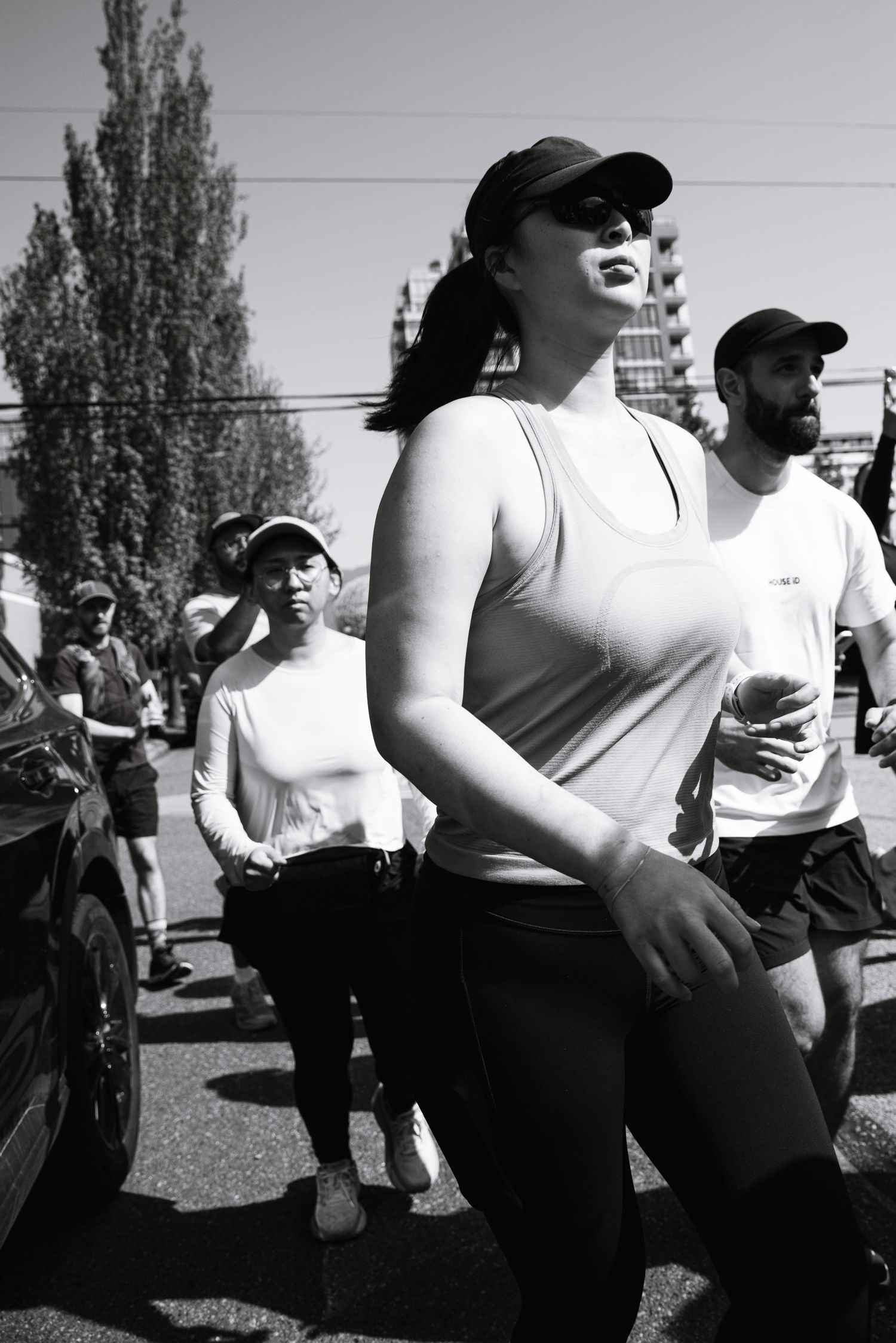 Black and white photo of runners in a race, woman in cap and tank top leading group on sunny street.