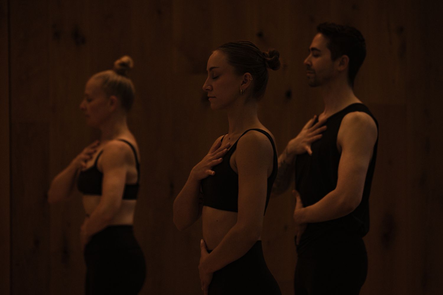 Three dancers in black attire stand in profile with hands on chests in a dimly lit wooden studio.