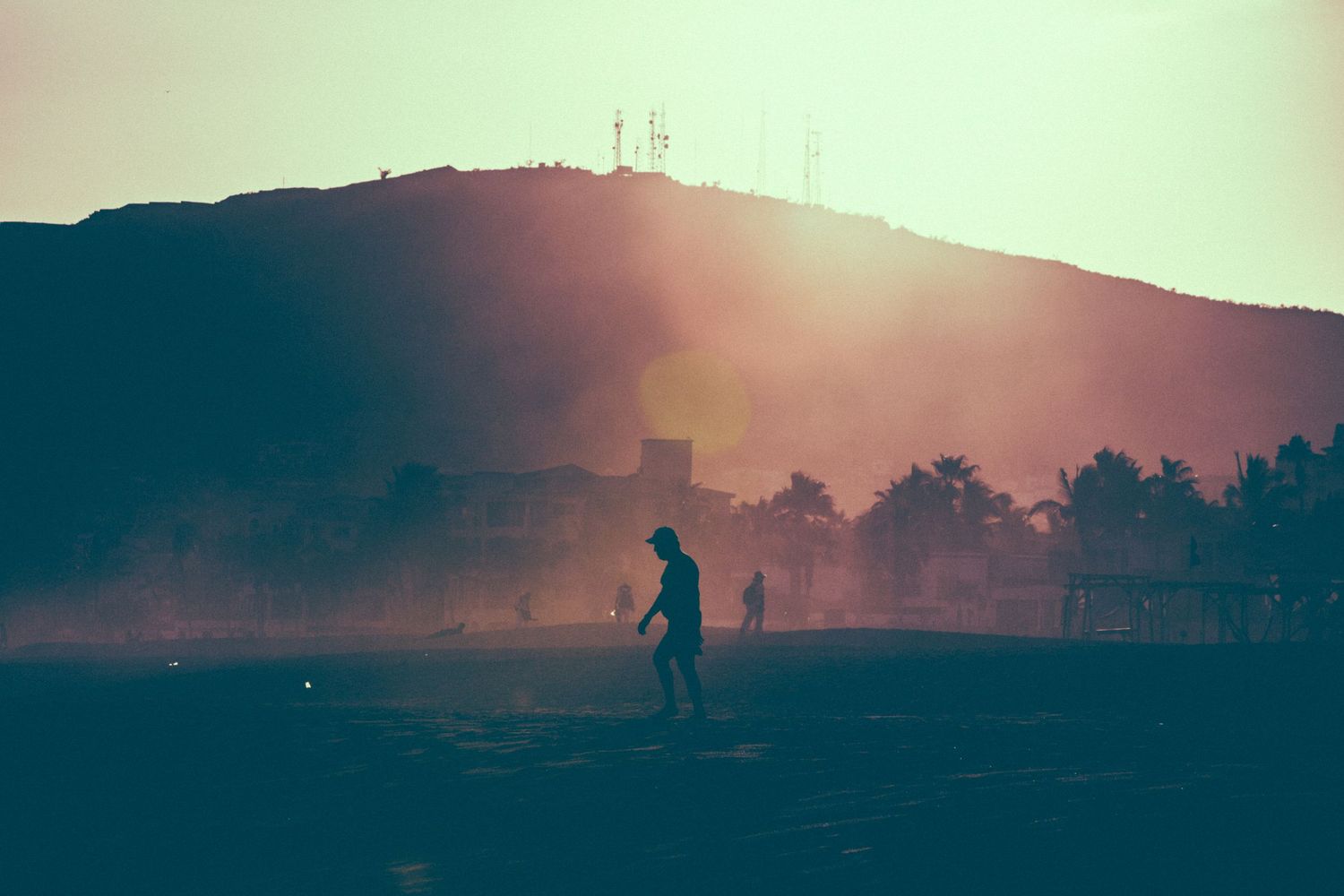 Silhouette of a person walking on a misty field at sunset with a hill and communication towers in the background.