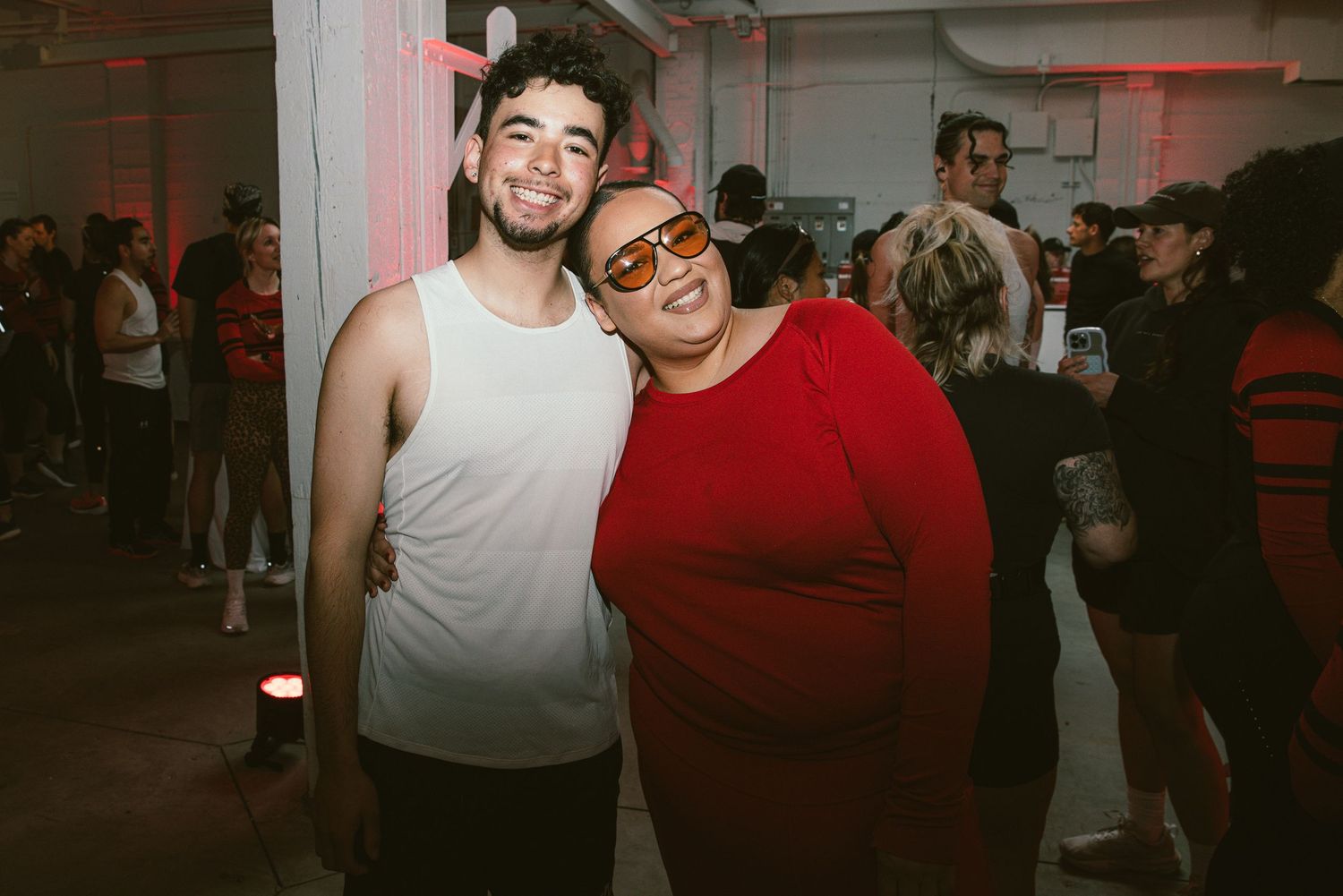 Two friends pose together at a lively indoor party, one in a white tank top and one in a red sweater.