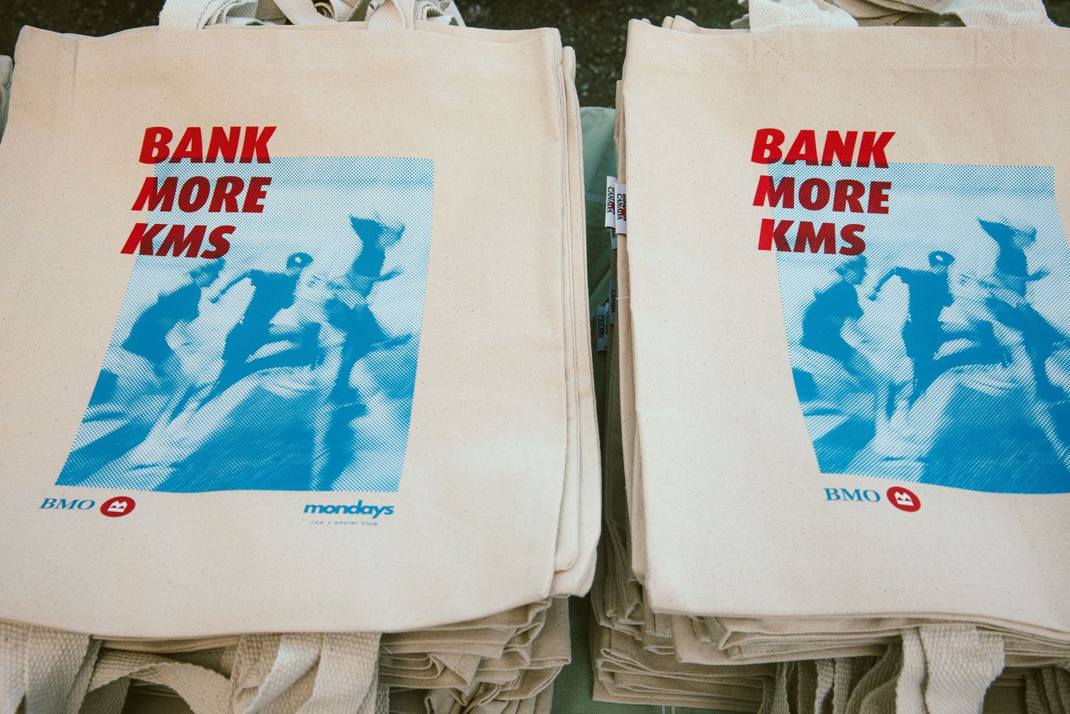 Tote bags printed with Bank More KMS slogan and blue graphic design, branded with BMO and Mondays logos.