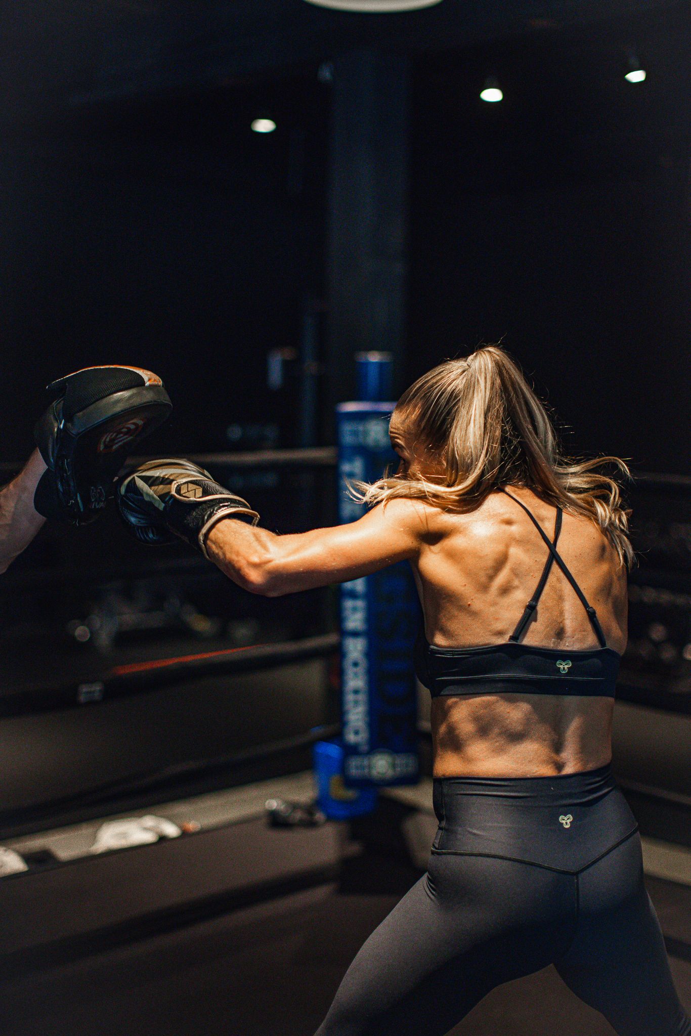Female boxer in sports bra throwing punch in dark boxing gym showing muscular back.