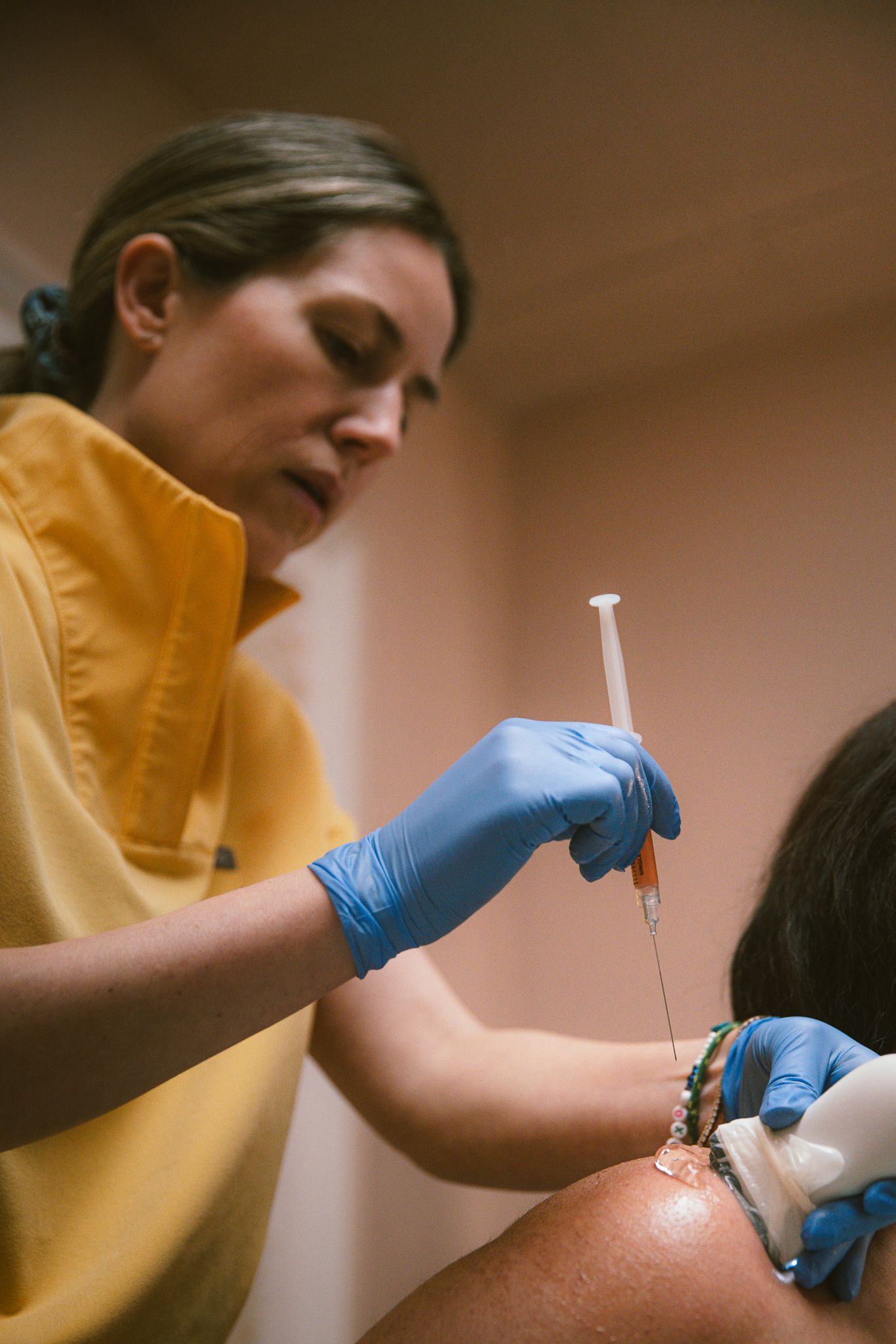 Healthcare professional in yellow scrubs administering an injection to a patient's shoulder while wearing blue latex gloves.