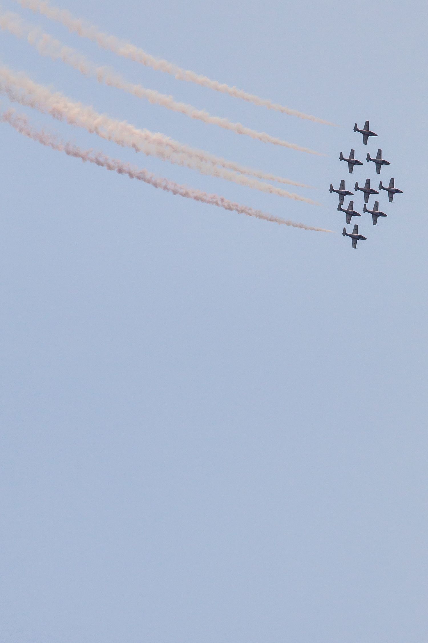 Seven aircraft perform a precise diagonal formation flight with trailing smoke trails against a clear blue sky.