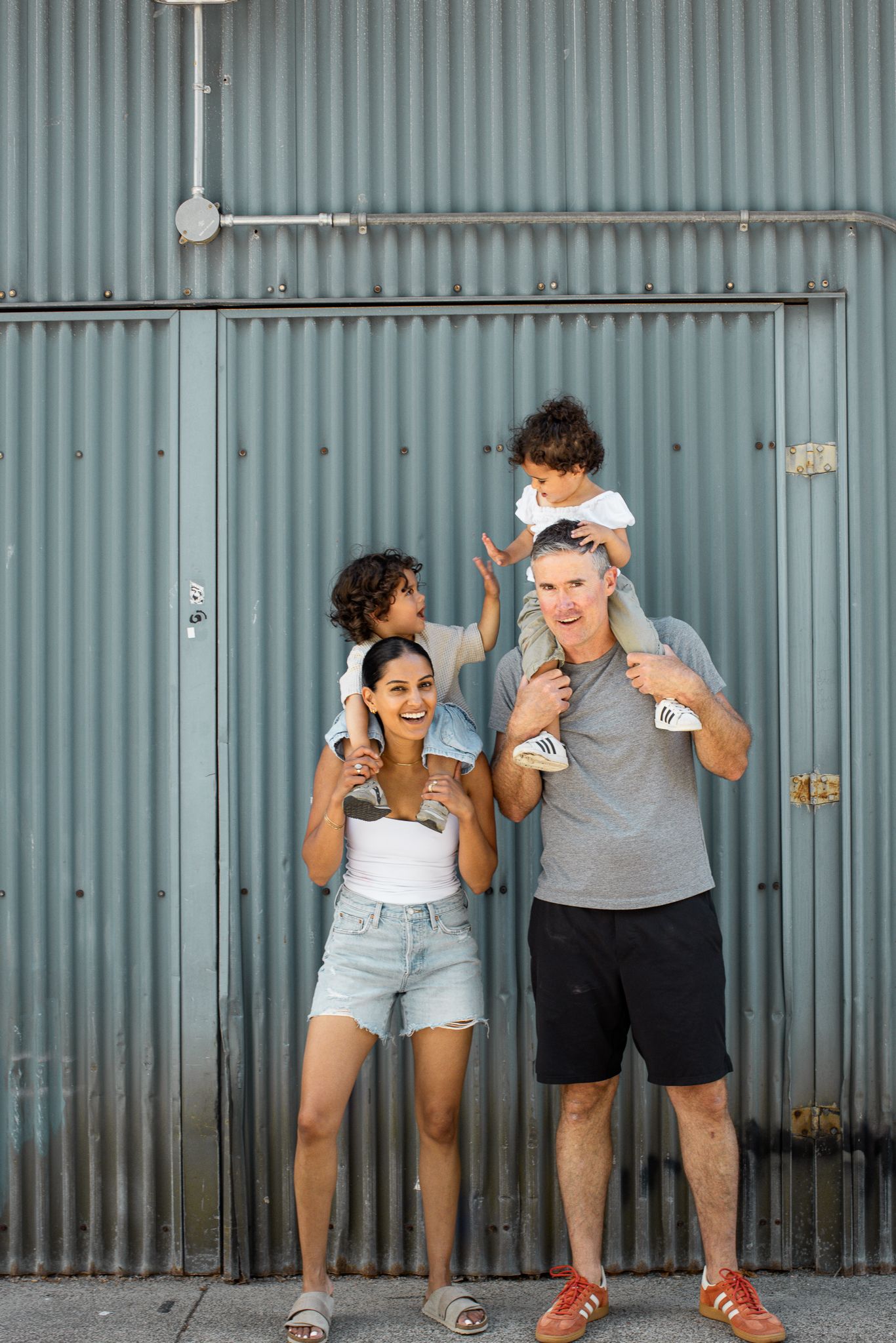 Happy family with two young children on their shoulders posing in front of a corrugated metal wall outdoors.