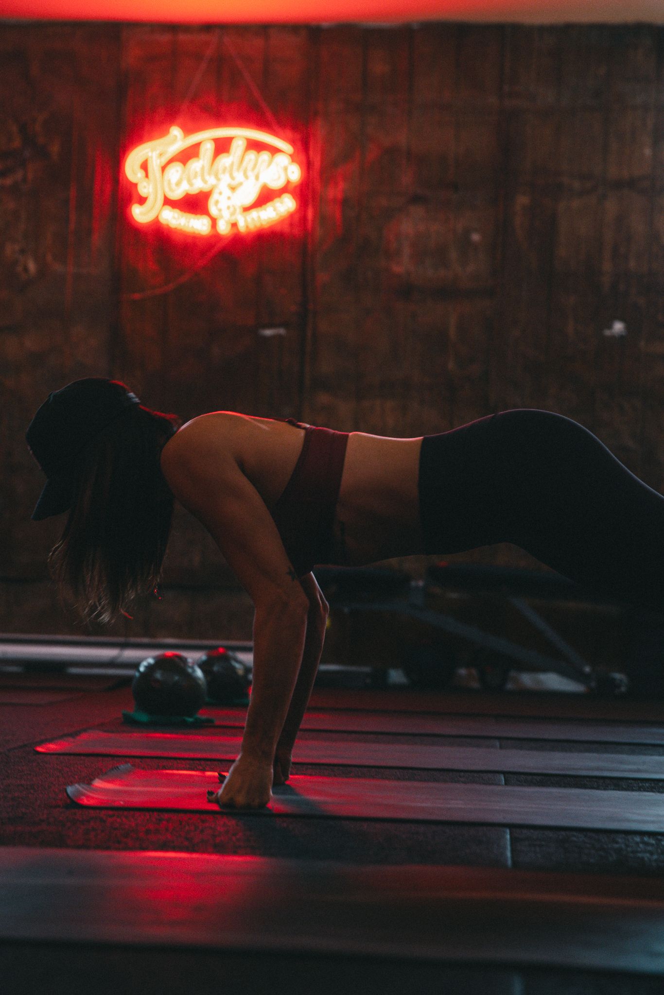 Woman performs a plank exercise on a mat in a dark gym lit by a red Teddy's Boxing neon sign.