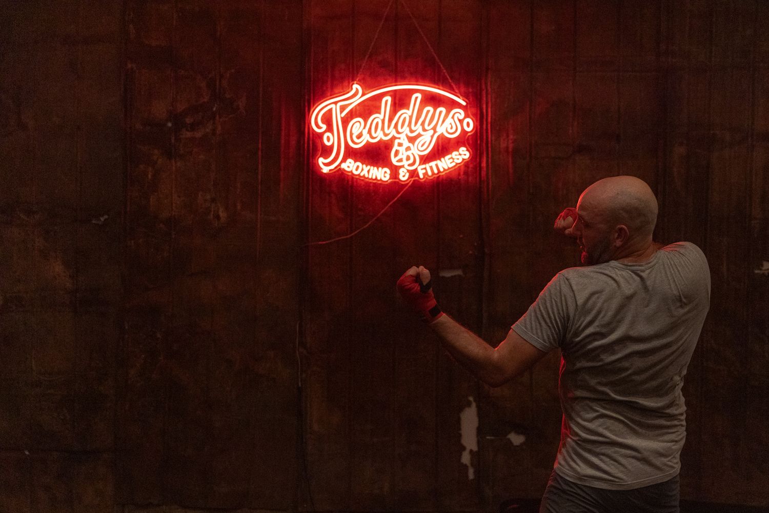 Man in boxing gloves punches toward a red neon Teddy's Boxing and Fitness sign on a dark wall.