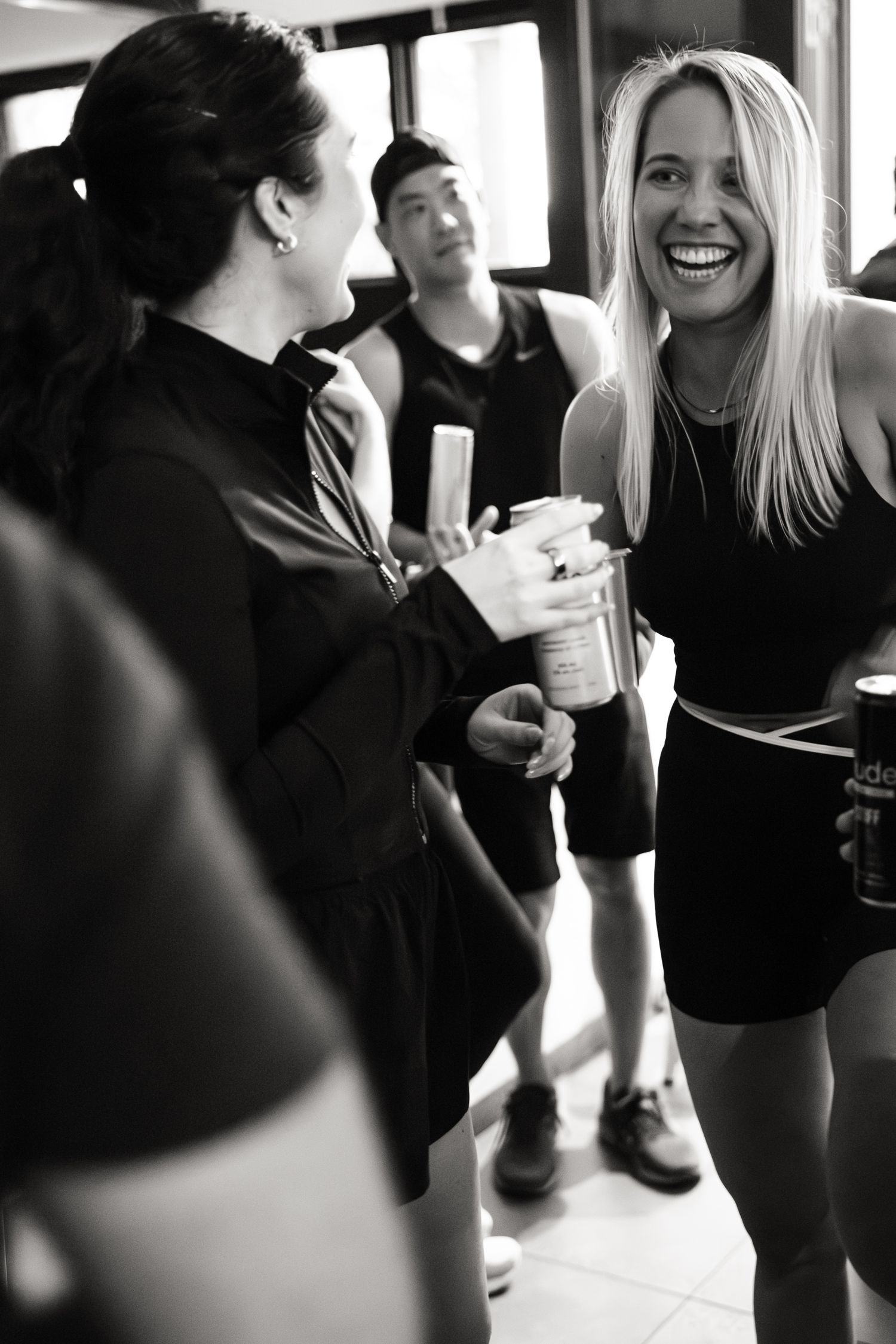 Two women laugh together at a post-workout social gathering, holding drinks in black and white photo.