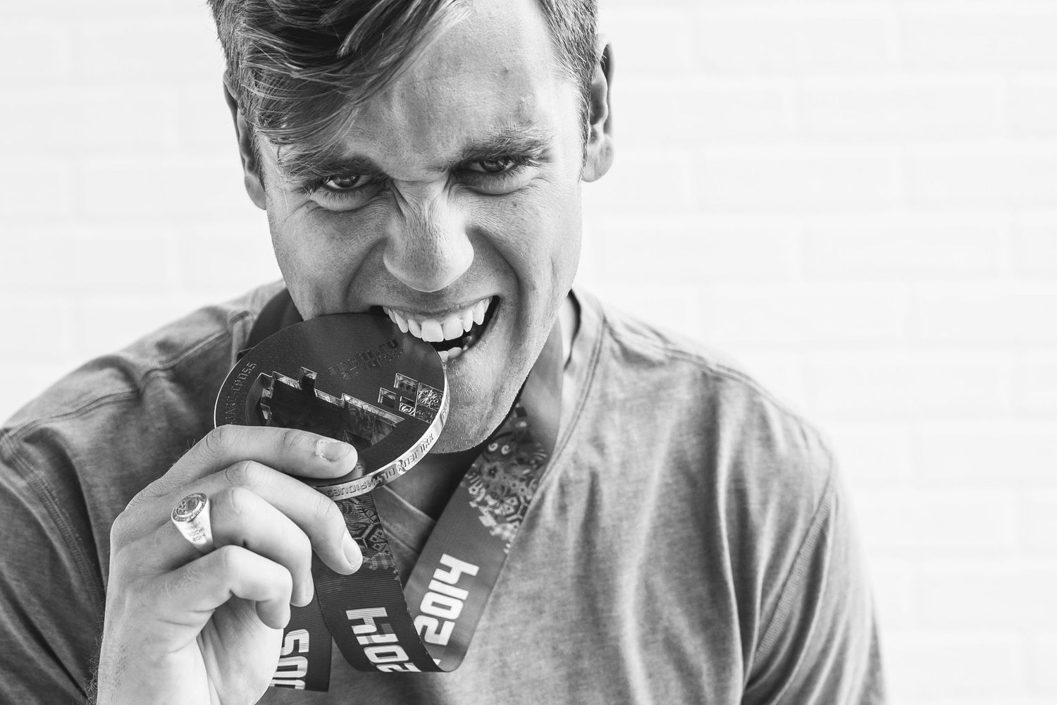 Black and white photo of a smiling man biting a medal around his neck, celebrating a race achievement.