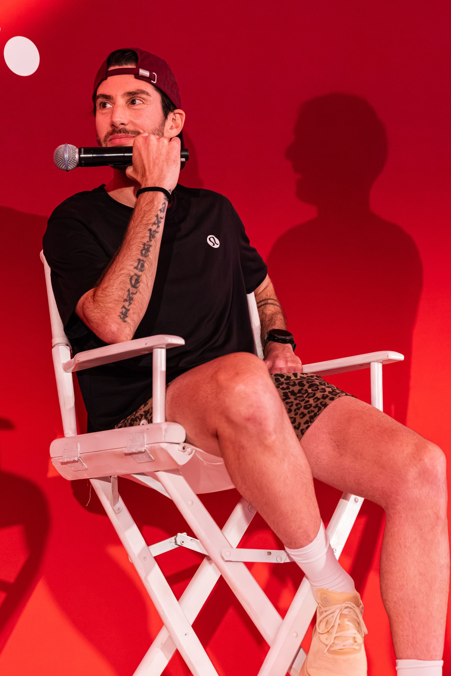 Tattooed man in black t-shirt and leopard print shorts sits in white director's chair holding microphone against red background.