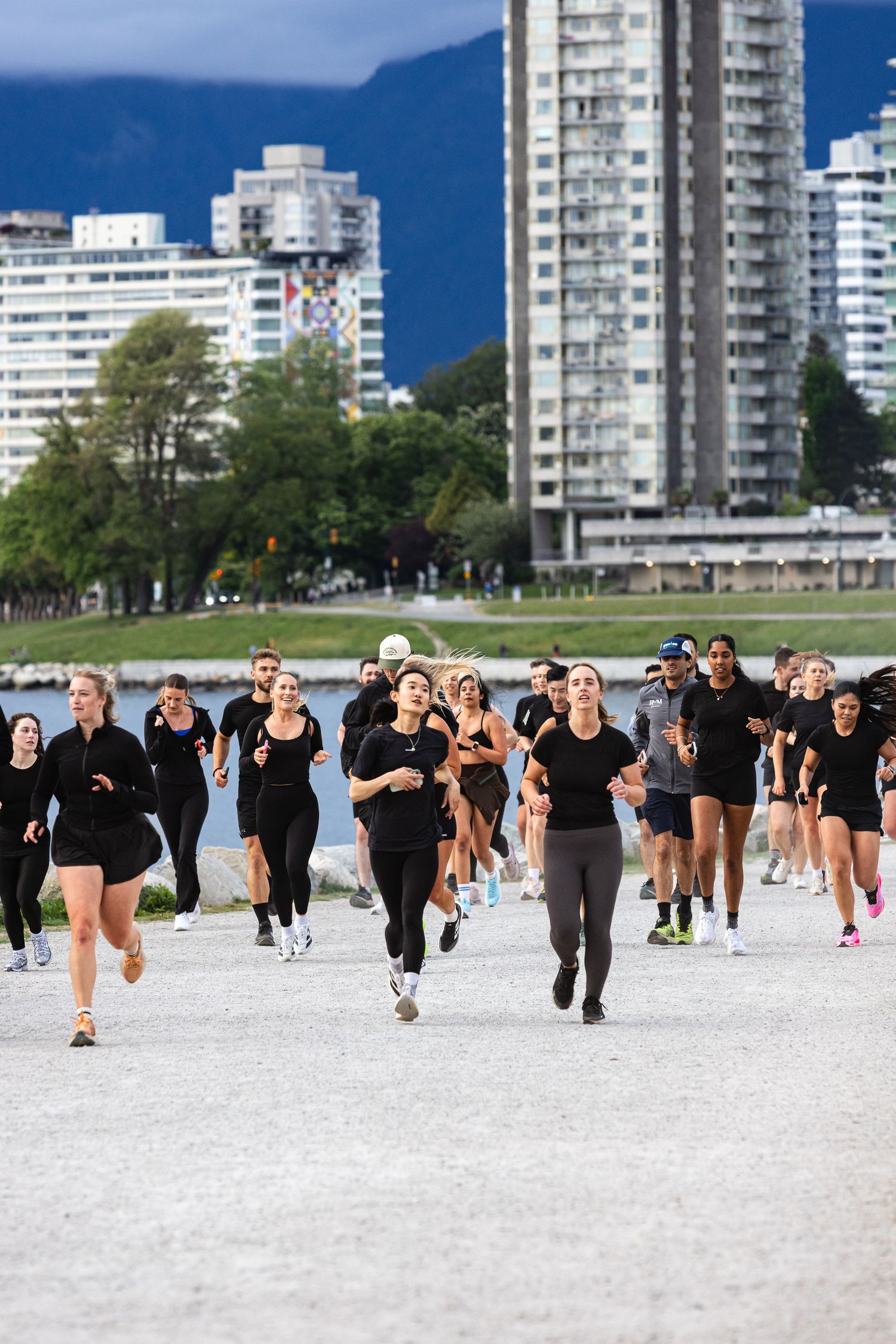 Large group of runners in black athletic wear sprint along a waterfront path with city skyline.