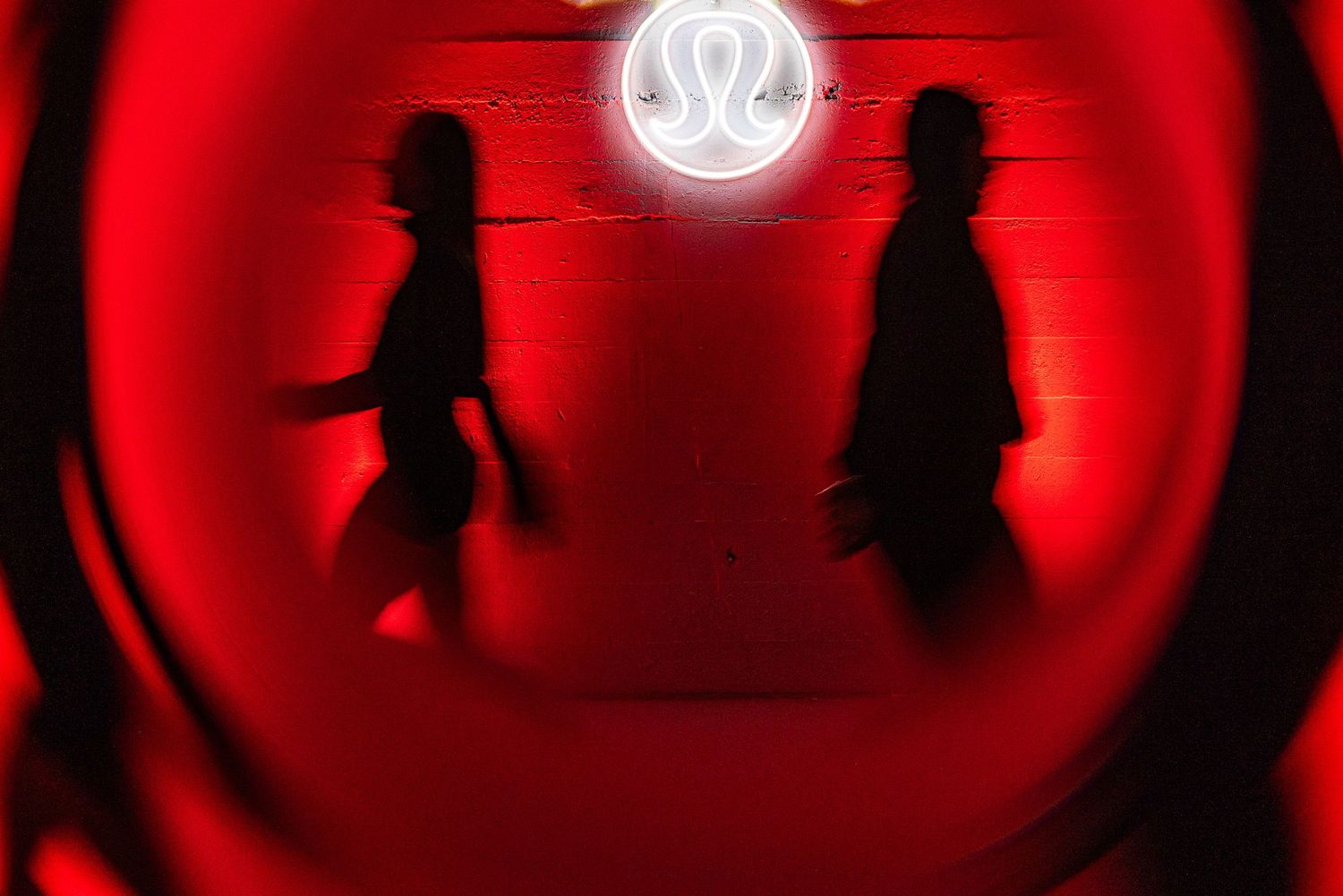Two silhouettes walking past a glowing lululemon neon logo sign on a red-lit wall.