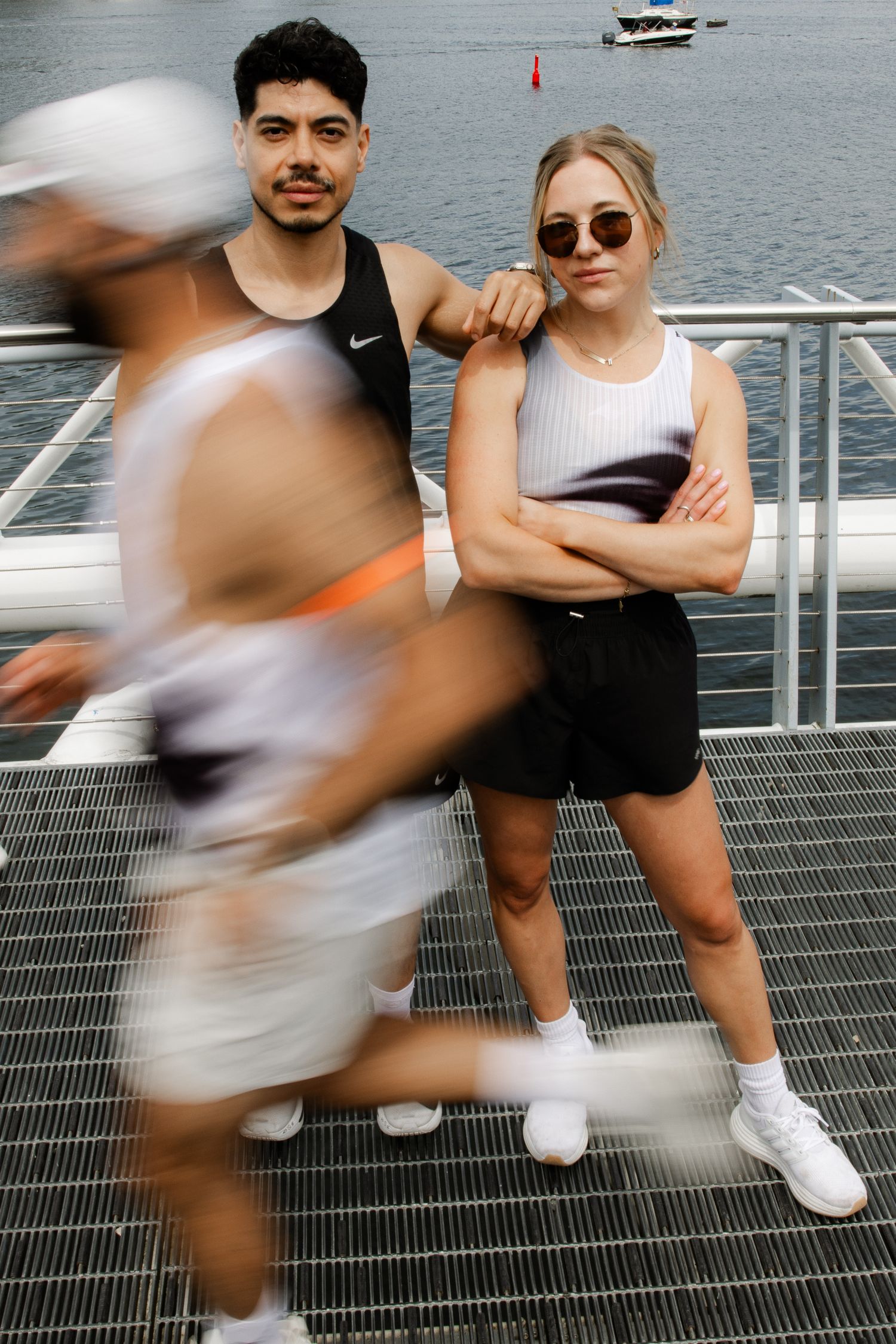Man and woman in athletic wear posing on waterfront bridge with blurred runner.