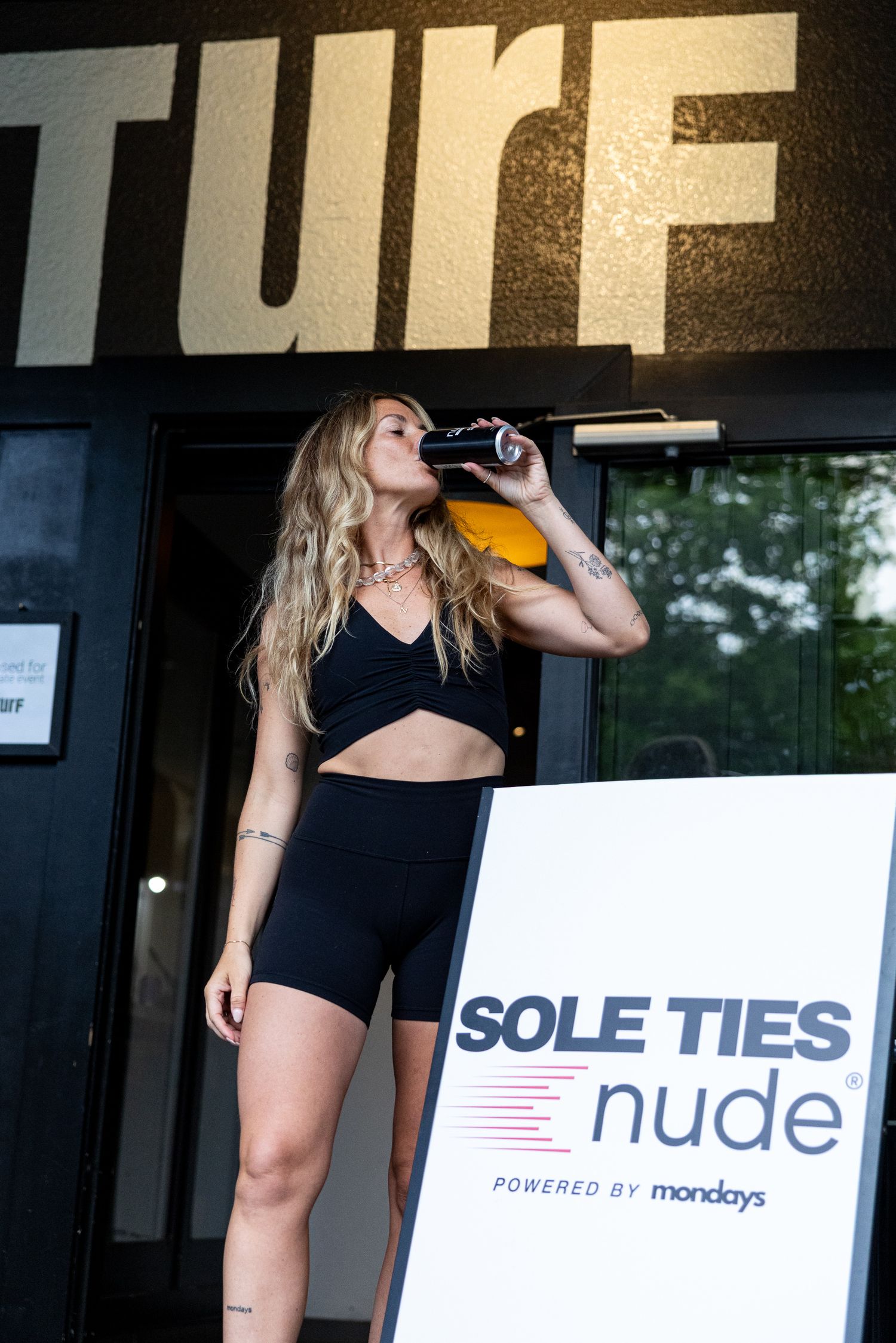 Woman in black athletic wear drinks from a can outside TURF gym near a Sole Ties Nude powered by Mondays sign.