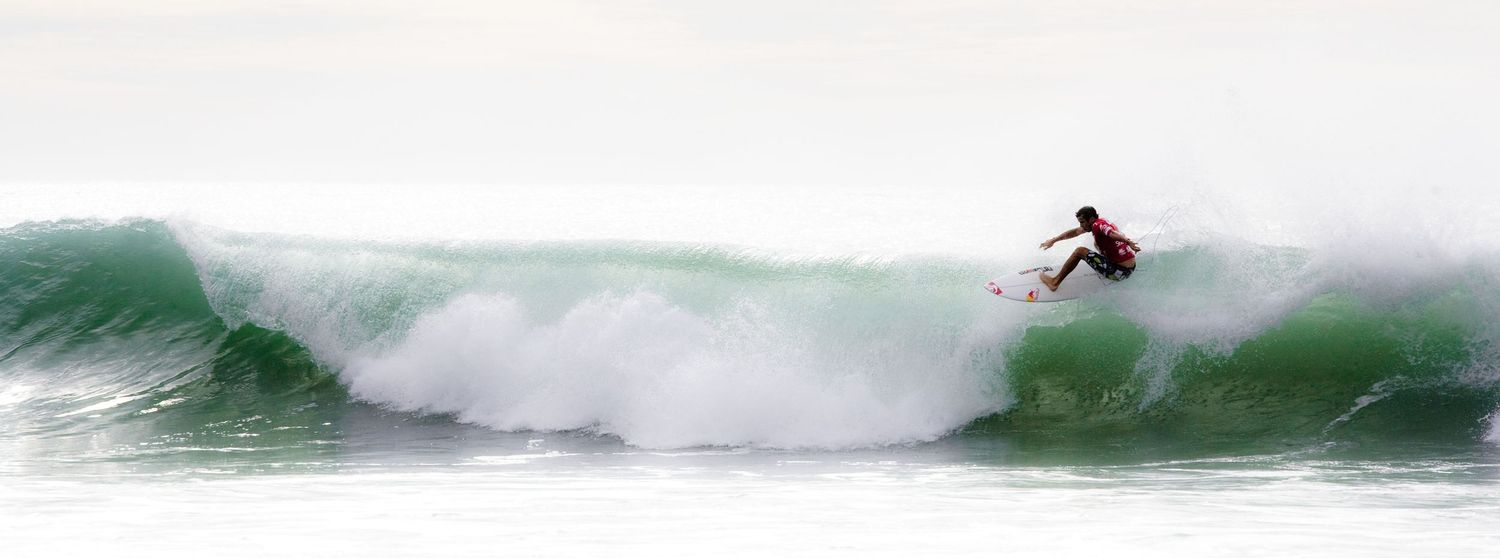 A surfer rides a large green wave in the ocean, performing an aerial maneuver against a misty white sky.