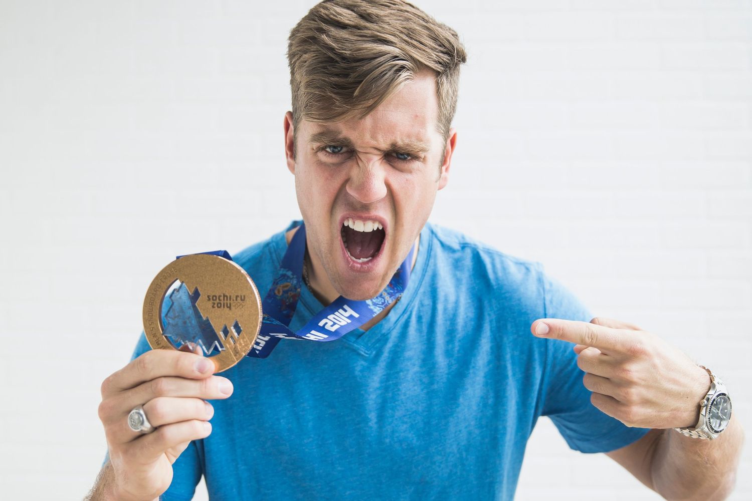 Excited young man in blue shirt pointing at gold medal around his neck, celebrating victory against white background.