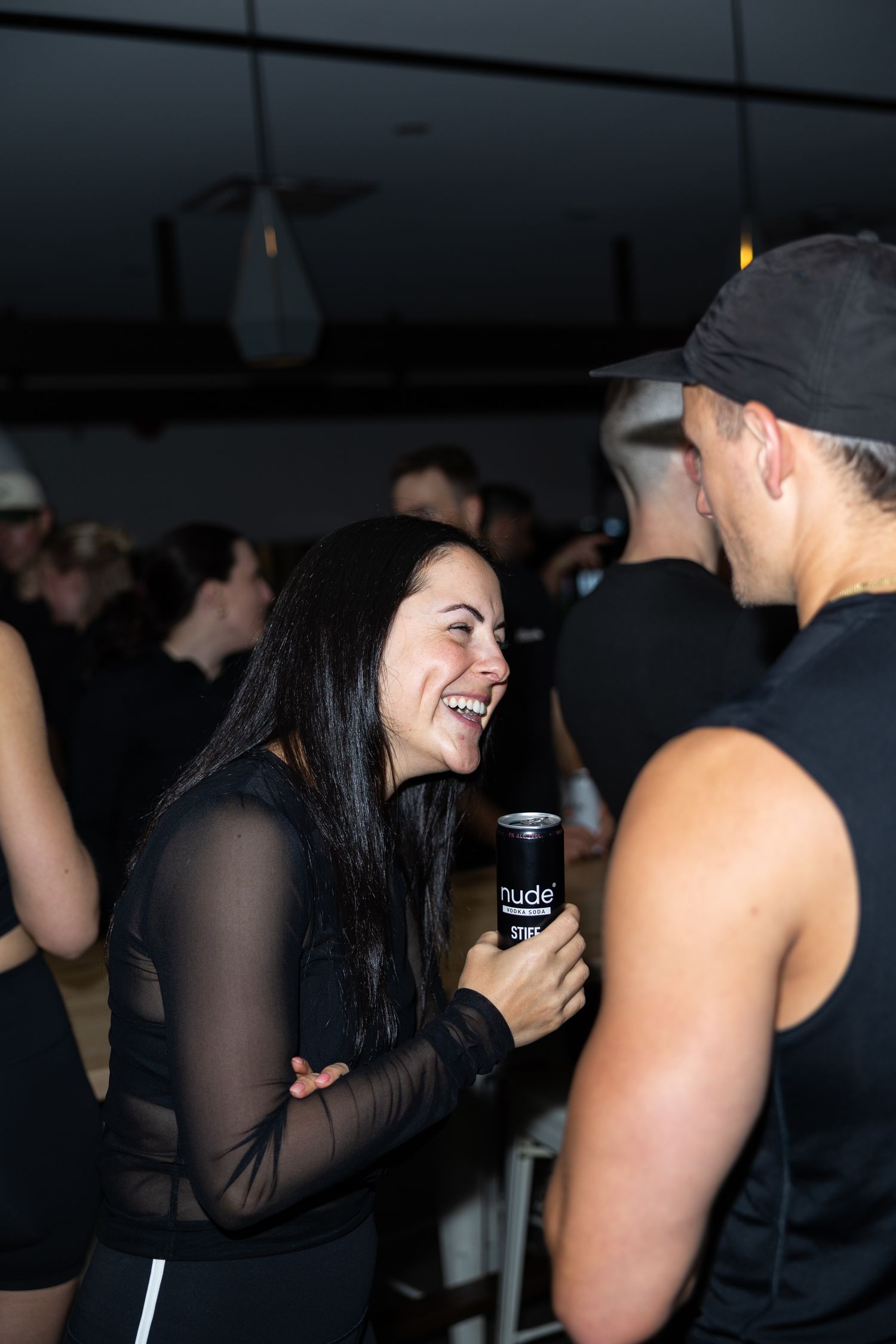 Woman laughing and holding a Nude drink can while socializing at a crowded indoor event, all dressed in black.