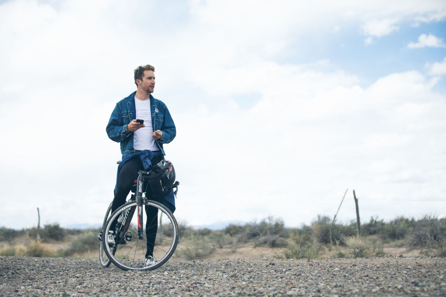 Man standing with bicycle on gravel road in open desert landscape, holding phone, wearing blue jacket on cloudy day.