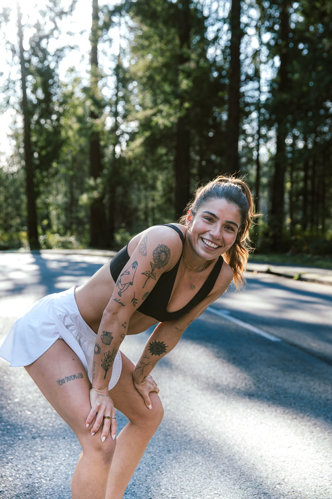 Smiling tattooed female athlete in black sports top and white shorts resting on a tree-lined road.