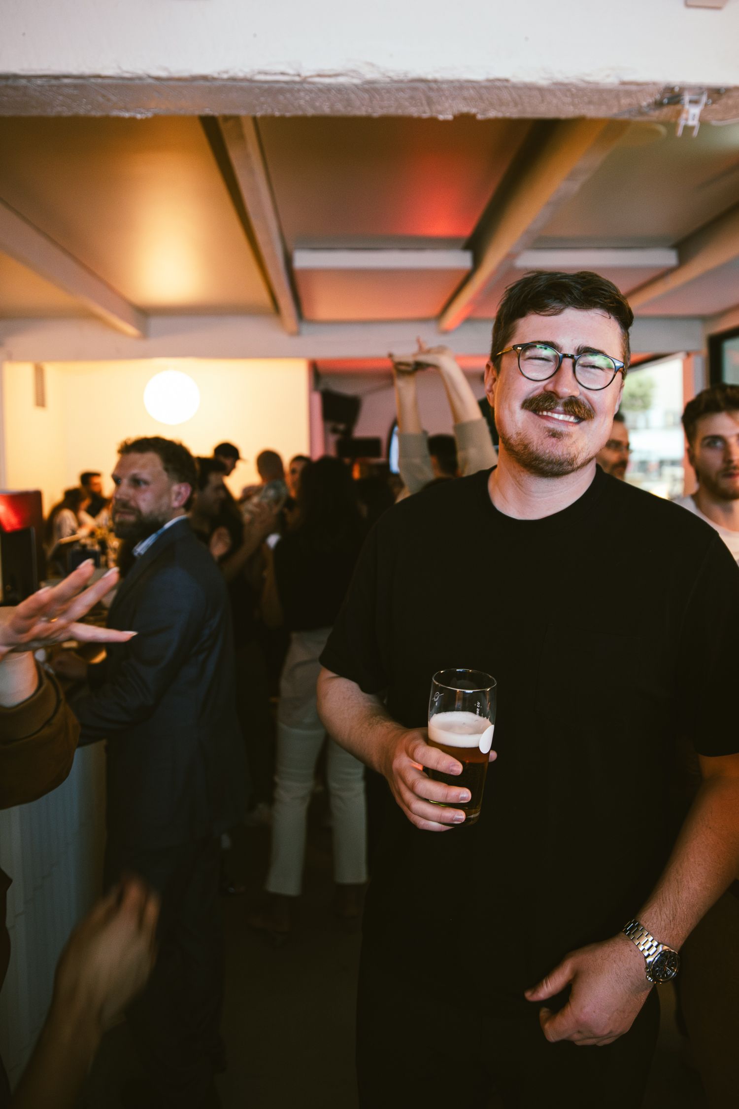 Smiling man with mustache and glasses holds a beer glass at a crowded indoor event.