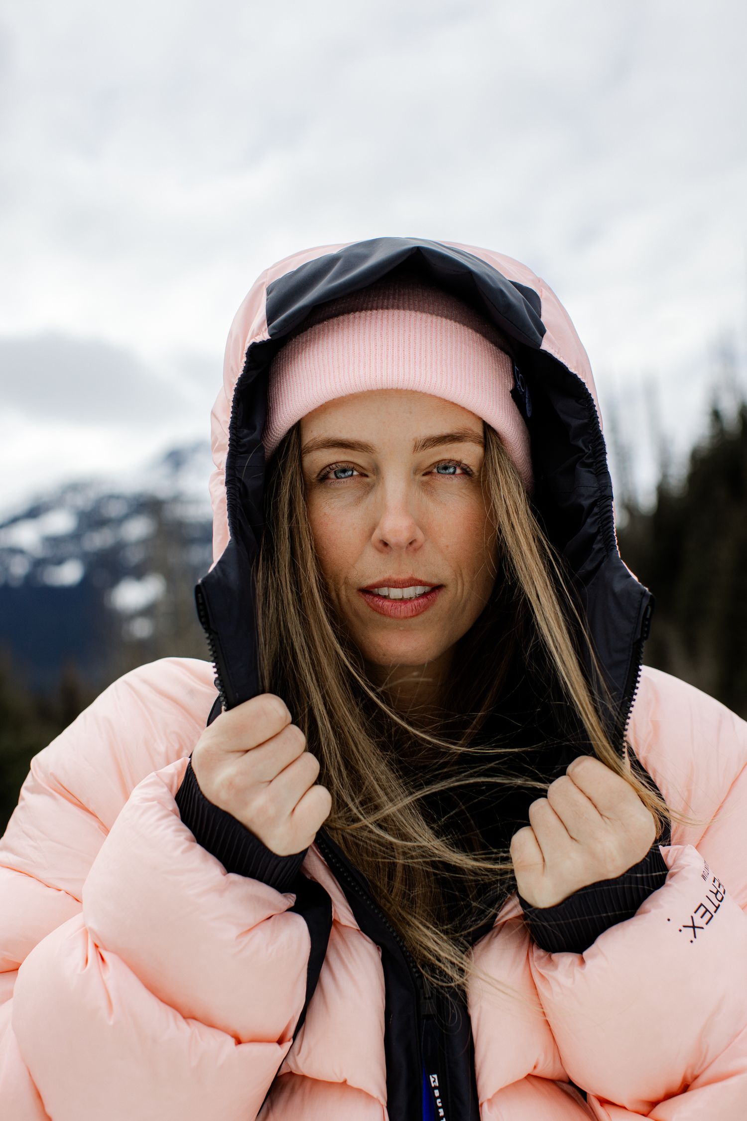 Portrait of woman with blue eyes wearing pink beanie and pink hooded puffer jacket with snowy mountains in background.