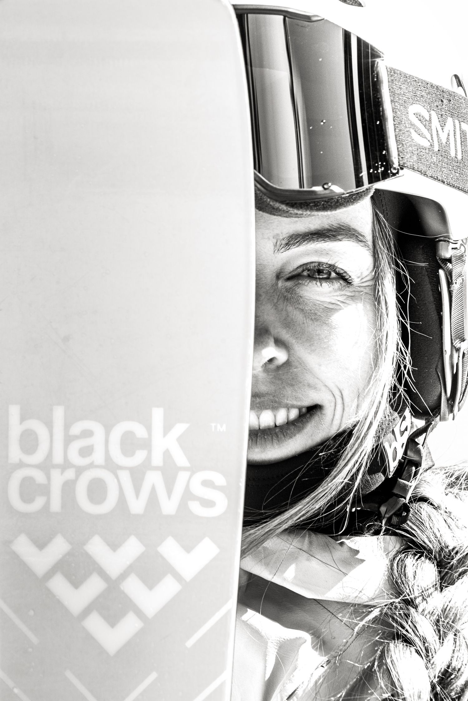 Black and white close-up of smiling female skier wearing Smith goggles and helmet beside a Black Crows ski logo.